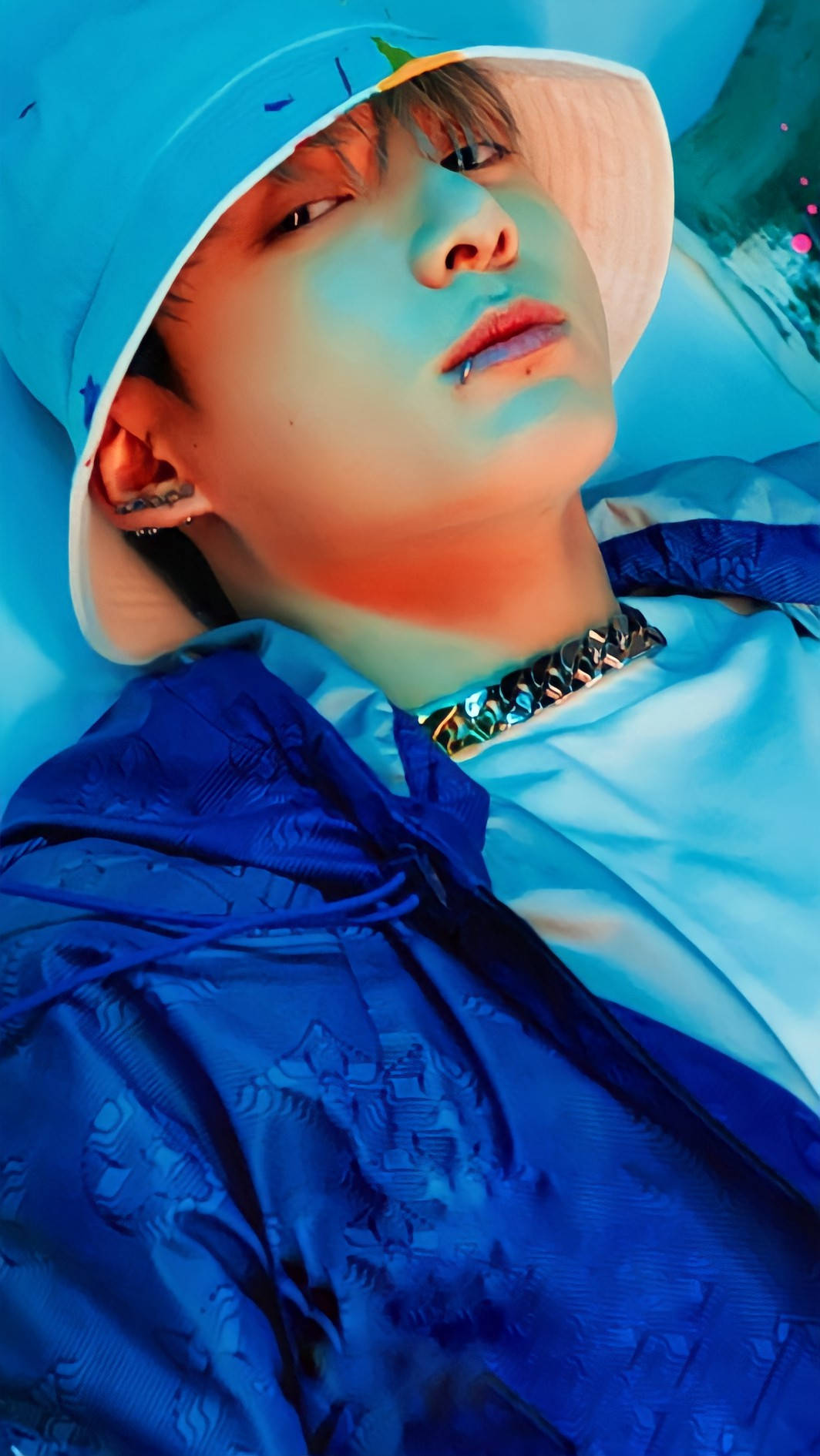 BTS JK迷人攝影 壁紙 — 一名戴著藍色漁夫帽和連帽衫的年輕男子，在霓虹藍光下戴著金屬鏈條項鍊向後躺。