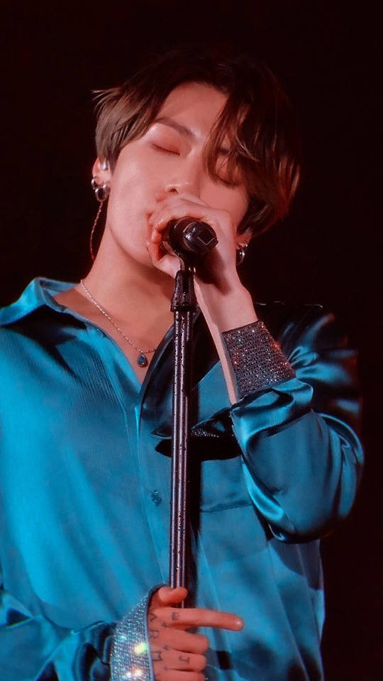 BTS Jungkook 唱歌 壁紙 — 一位表演者閉著眼睛對著麥克風唱歌，身穿閃亮的青綠色緞面襯衫，在舞台燈光下。