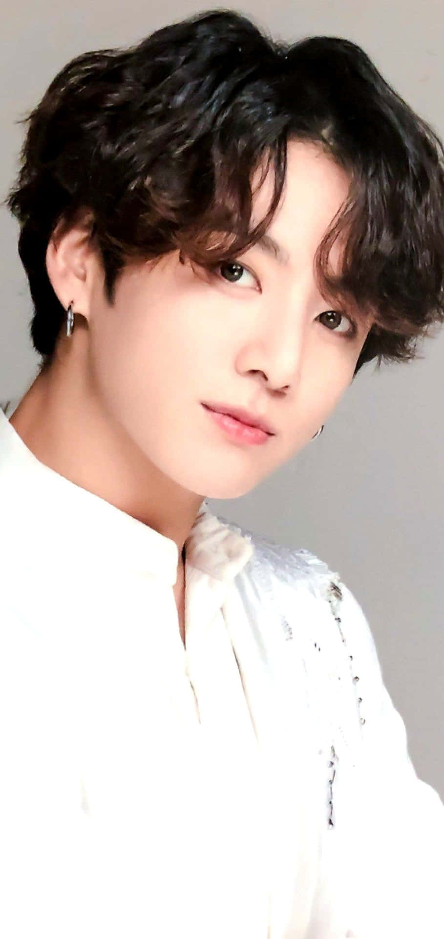 BTS Jungkook展現冷酷姿態 壁紙 — 年輕男子特寫肖像，深色捲髮，戴銀色耳環，身穿白色質感襯衫，背景為純灰色。