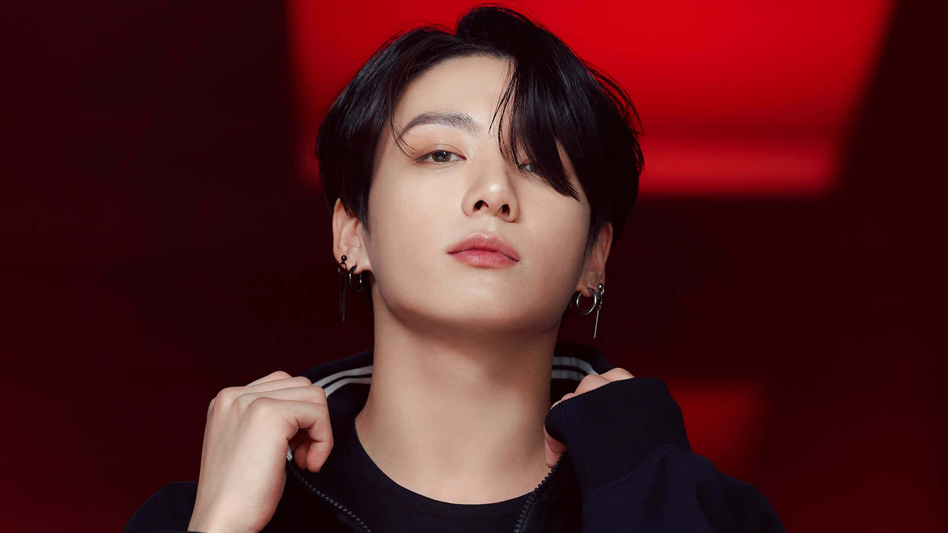 BTS Jungkook攝影寫真 壁紙 — 一位黑髮戴著圓環耳環的年輕男子，在深紅色模糊背景前向上凝視，同時調整他的黑色連帽衫。