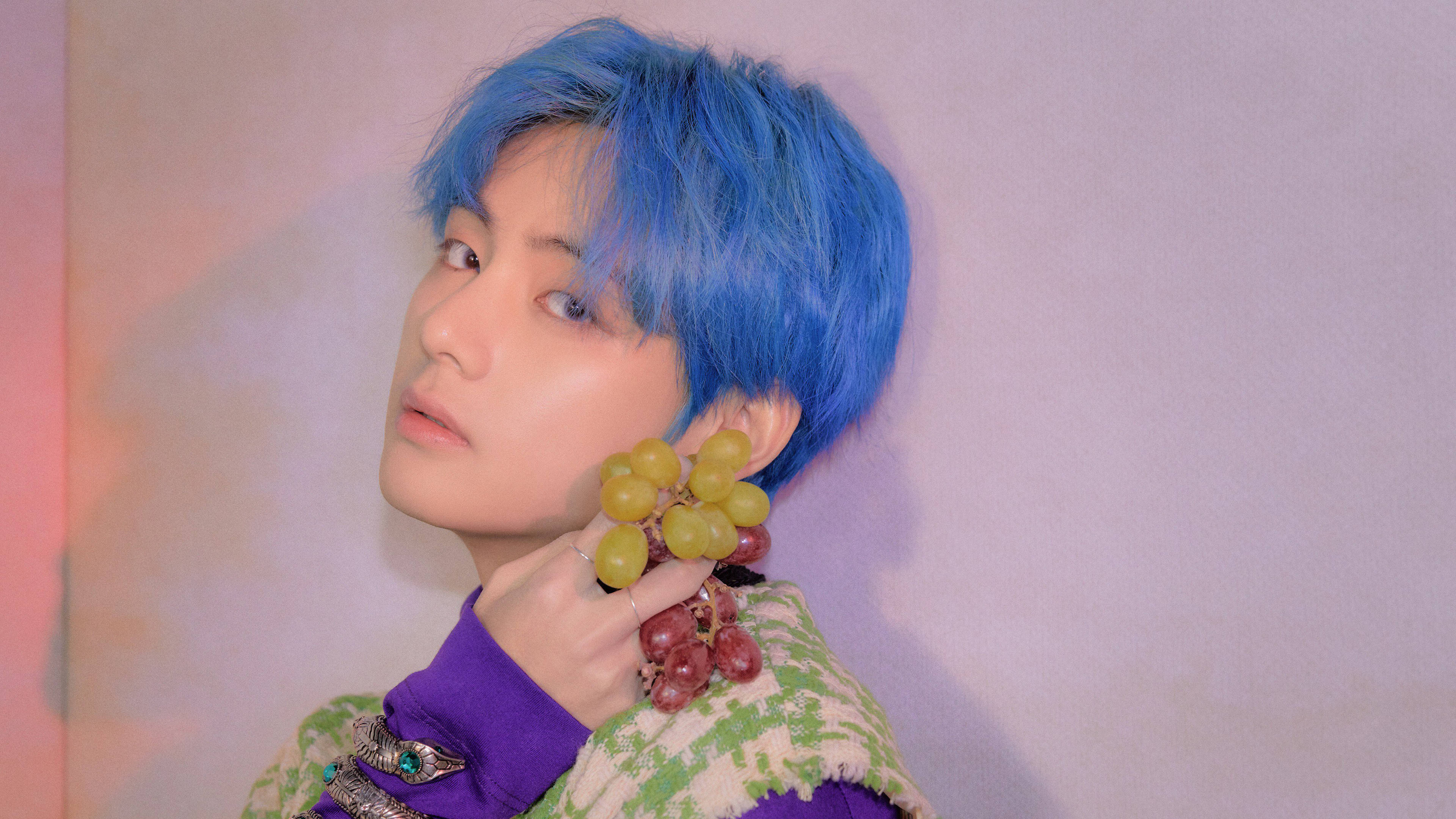 BTS 田柾國 手持葡萄 壁紙 — 一個擁有鮮豔藍色頭髮的人，手持一串綠色和紅色的葡萄，背景是柔和的粉彩漸變牆。