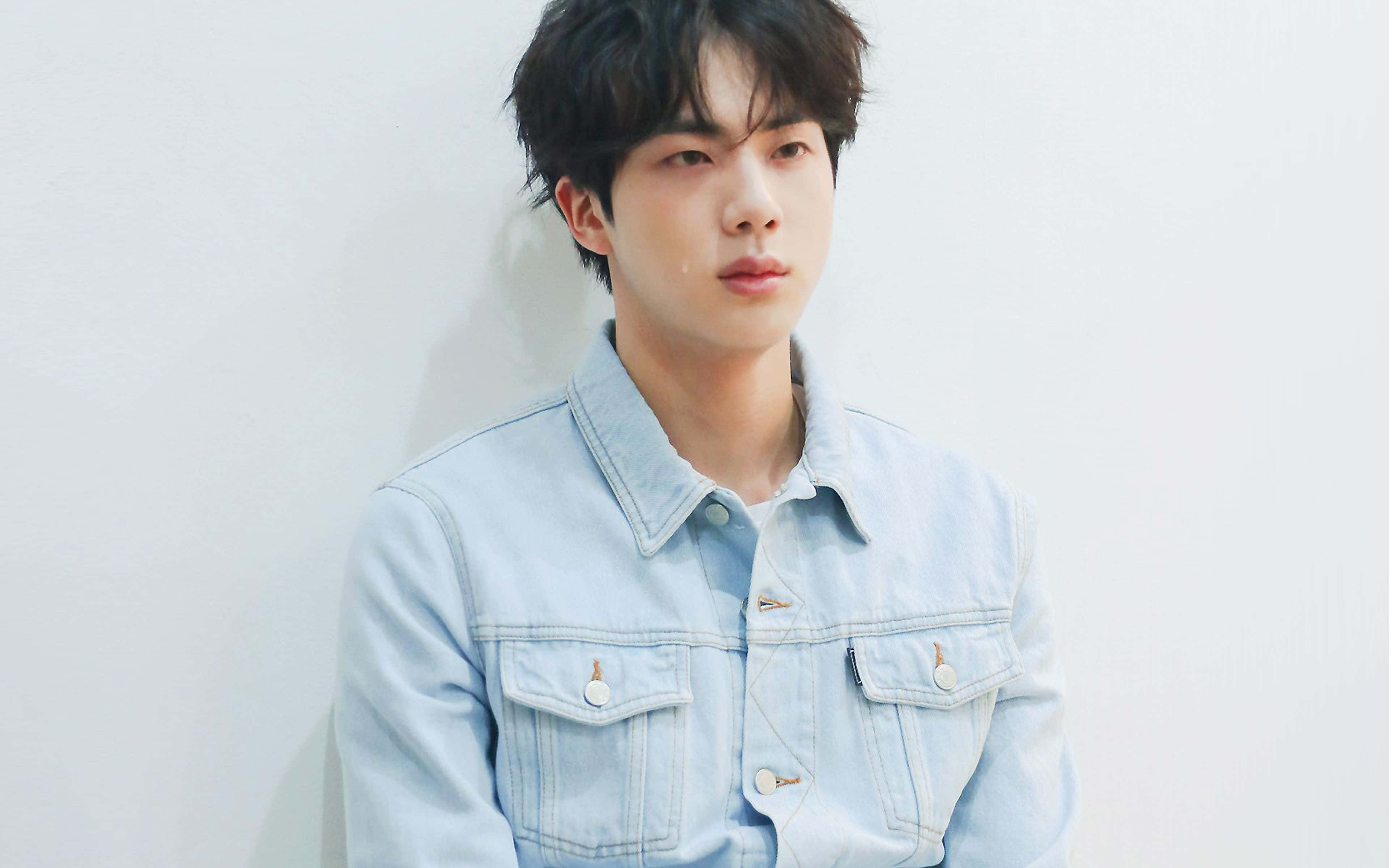 BTS Mac，工作與娛樂的完美夥伴 壁紙 — 一位穿著淺藍色牛仔外套的年輕男子，在柔和自然光線下靠著白色牆壁。