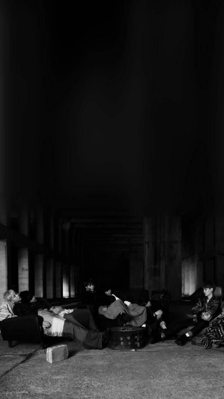 BTS 黑色休息時刻壁紙 壁紙 — 一群人在昏暗的柱廊走廊中休息，黑白風格，帶有戲劇性的陰影和極少的光源。