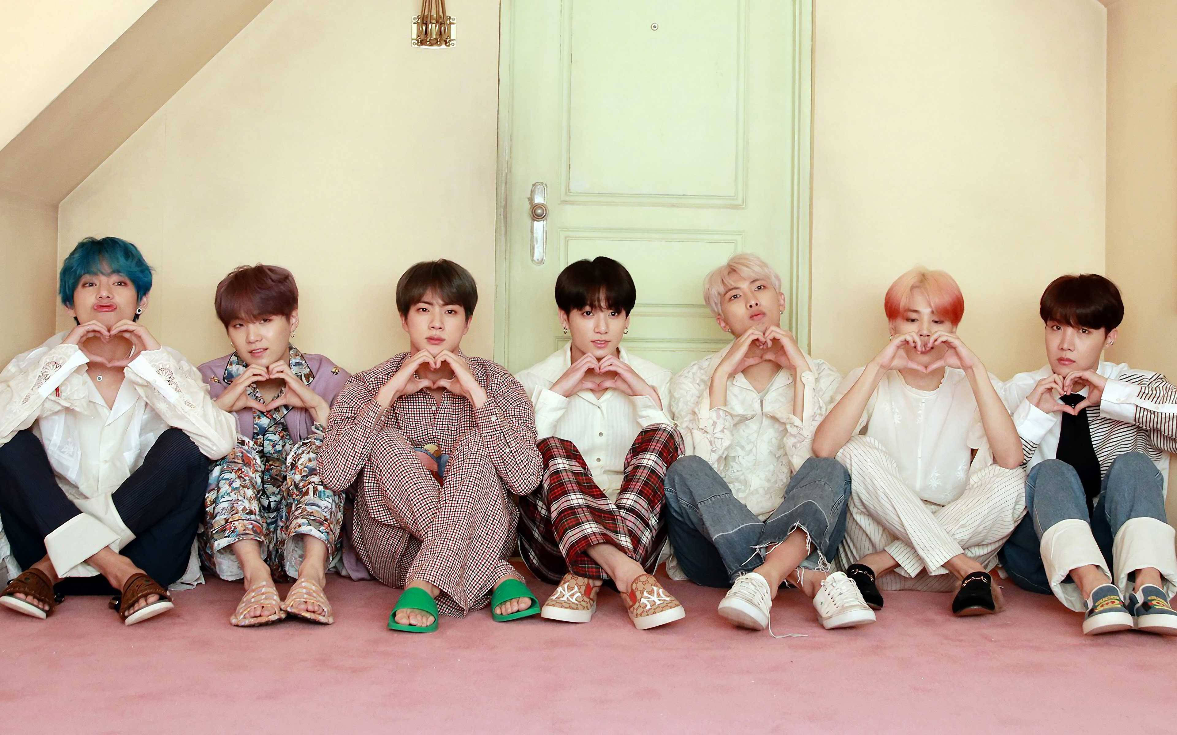 BTS成員門前比心桌面壁紙 壁紙 — 七位年輕男子擁有色彩繽紛的頭髮，坐在粉紅色地板上，對著一扇粉綠色門用手做出心形。