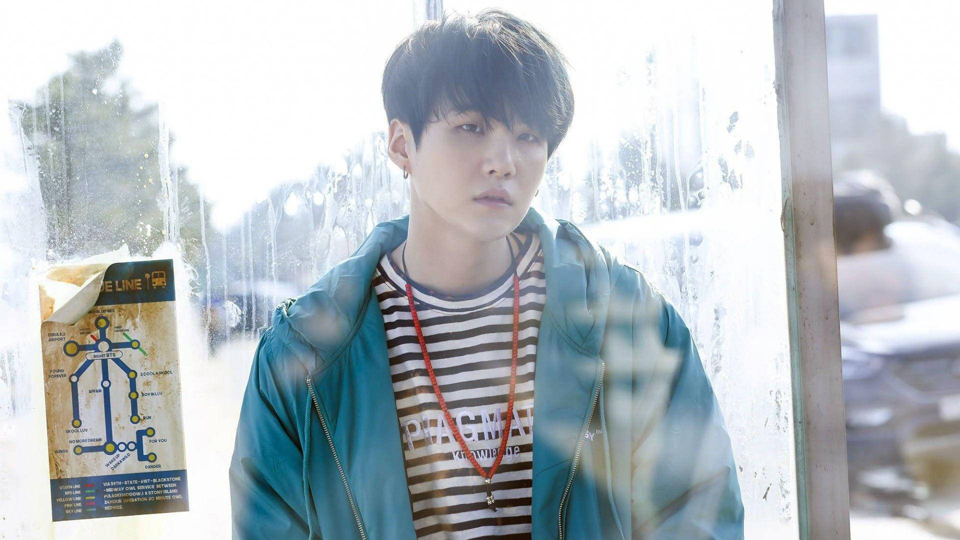 休閒風 BTS Suga 壁紙 — 一位身穿青綠色外套和條紋襯衫的年輕男子站在有雨痕的窗邊，旁邊有一張地鐵路線圖海報。