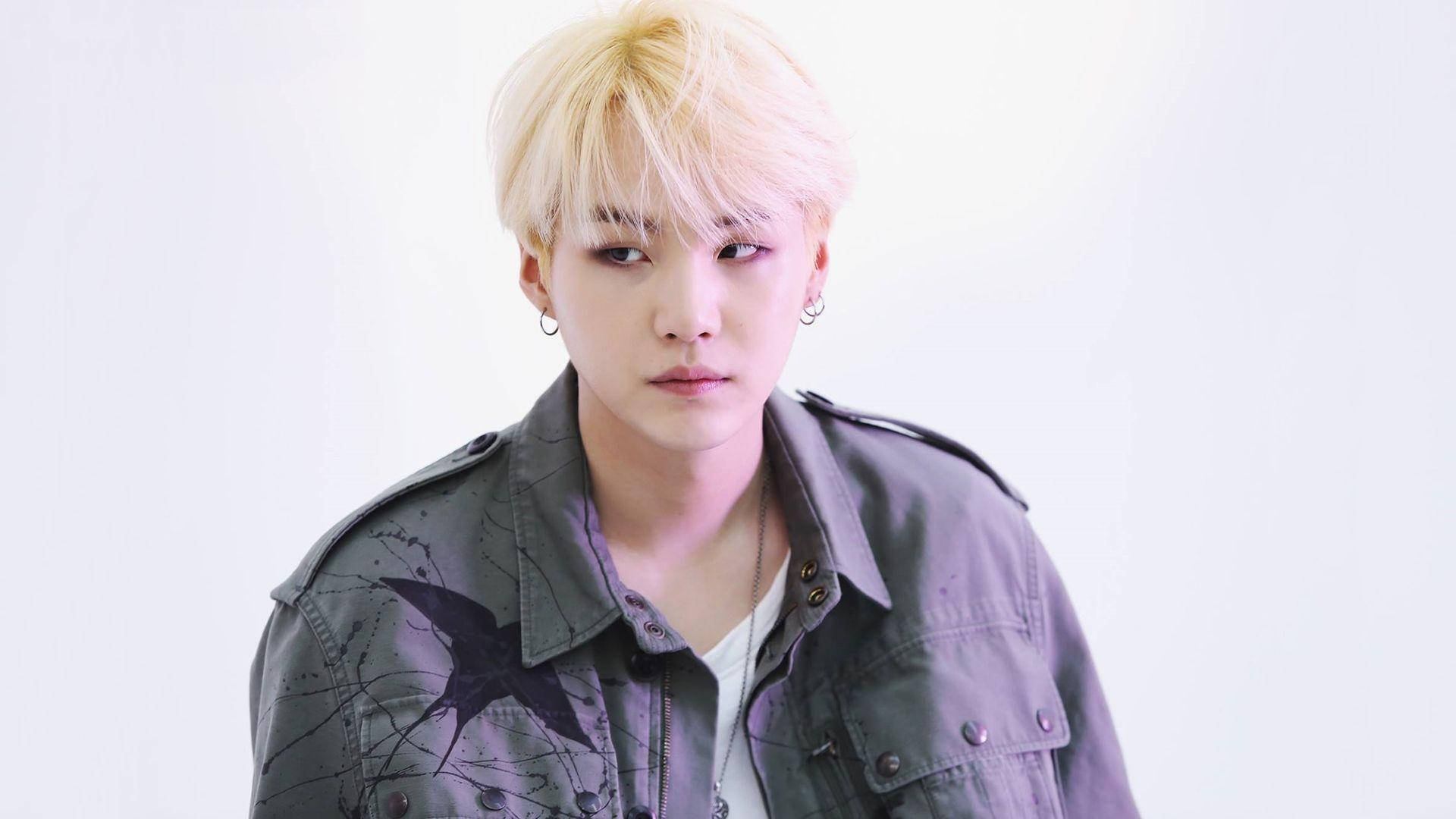 嚴肅金髮 BTS Suga 壁紙 — 一位年輕男子，擁有鉑金色頭髮和深色星星圖案外套，在純白背景前凝視著。