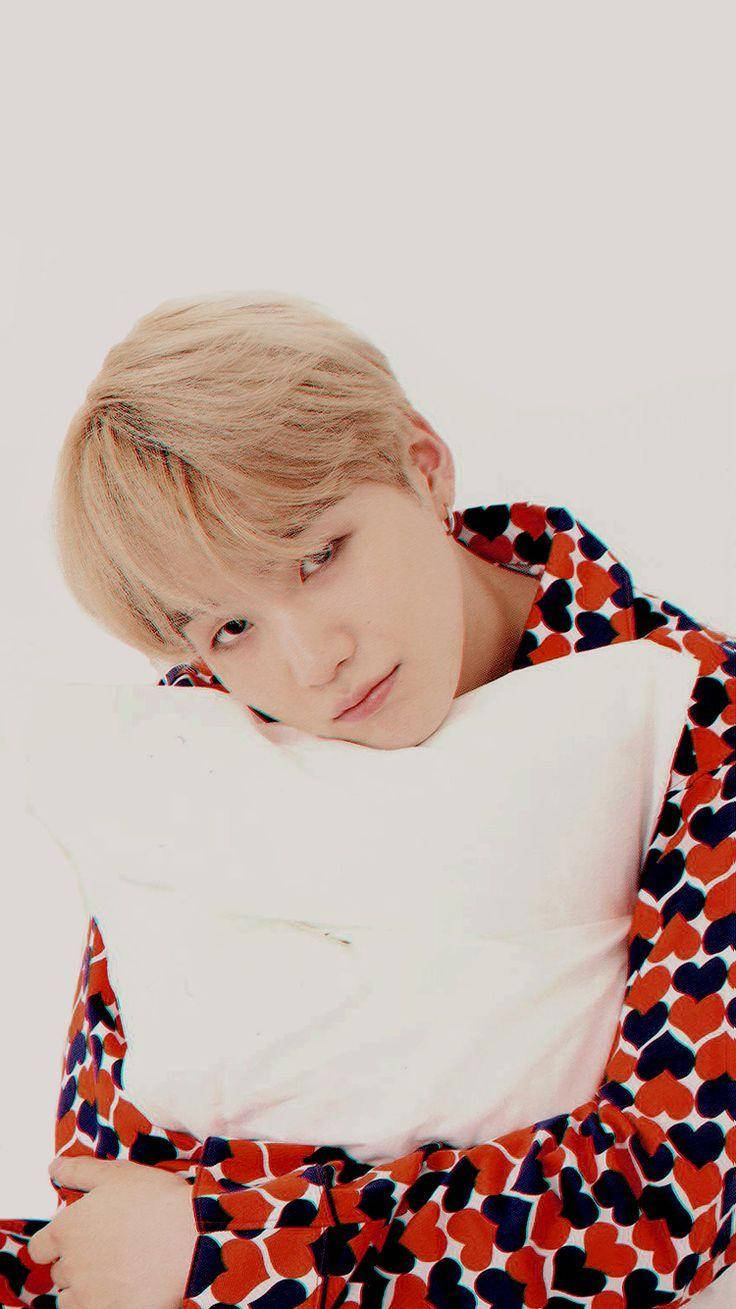 BTS Suga 睡衣造型 壁紙 — 一位金髮年輕男子將頭靠在白色枕頭上，身穿紅藍心形圖案睡袍，背景為純白色。