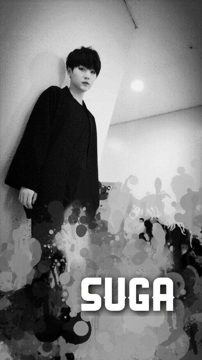 BTS Suga 黑白影像 壁紙 — 一個黑白肖像，一個穿著深色外套的年輕男子靠著牆，帶有抽象墨水飛濺和下方粗體文字。