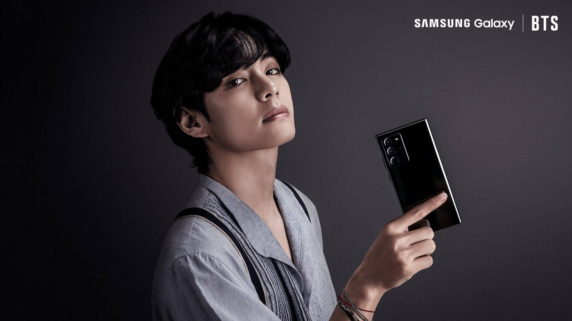 BTS Taehyung Samsung Galaxy 廣告 壁紙 — 一位穿著灰色襯衫的年輕男子在深灰色工作室背景前拿著一部黑色Samsung Galaxy手機。