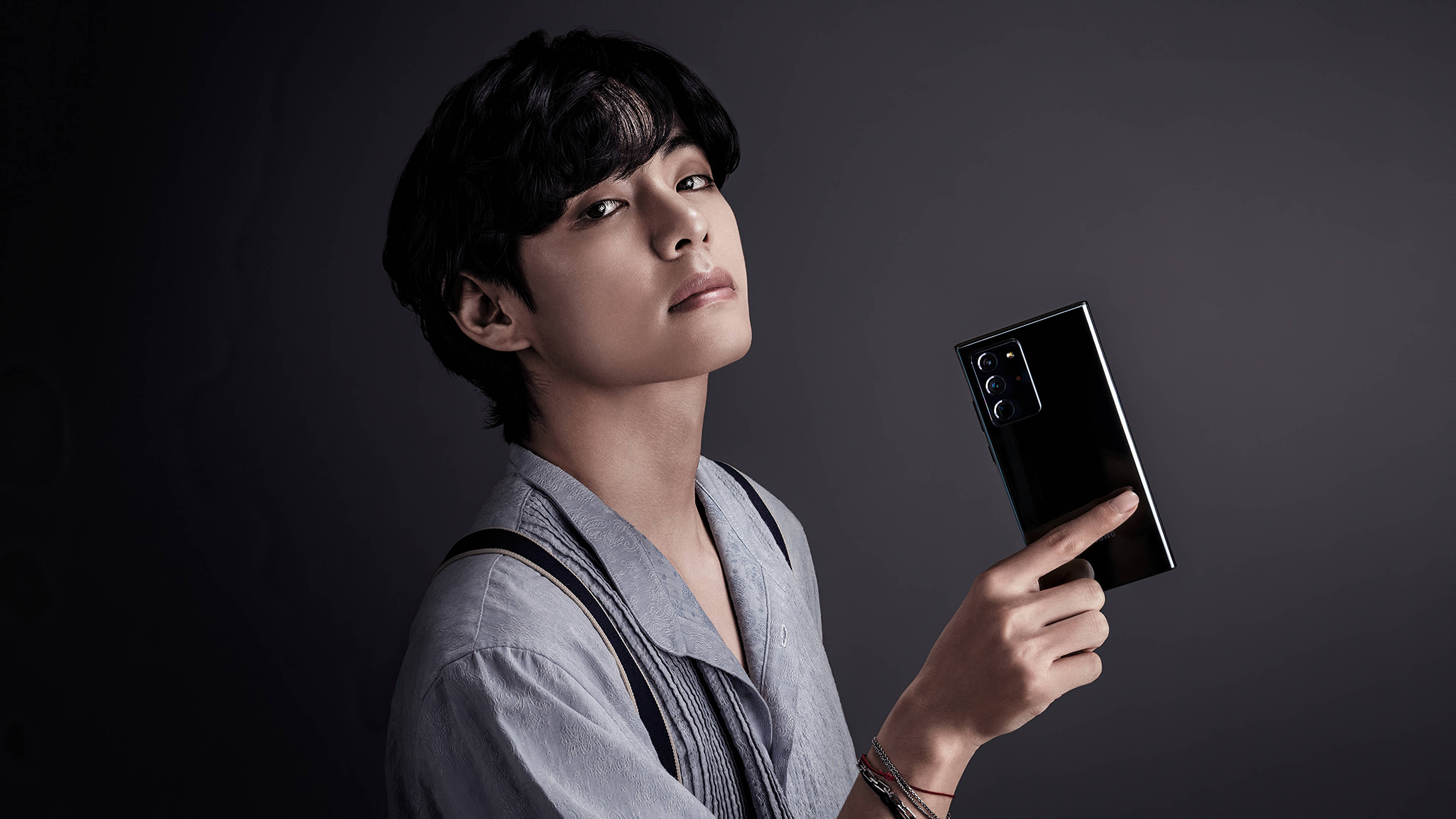 BTS V 擁抱三星手機 壁紙 — 一位身穿灰色襯衫的年輕男子在深灰色工作室背景前拿著一部黑色智能手機。