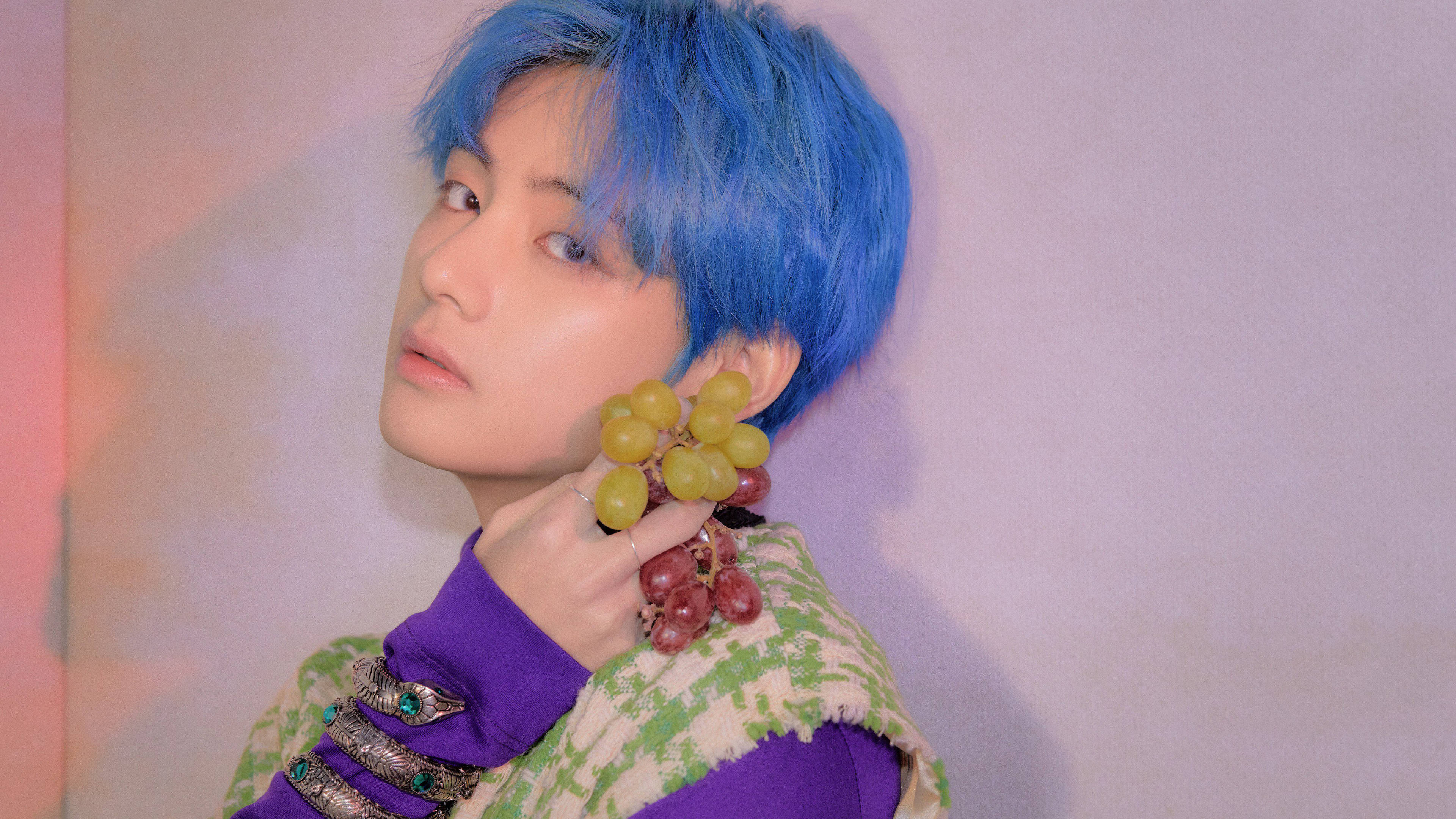 BTS V 手持葡萄 壁紙 — 一個擁有鮮豔藍色頭髮的人，手持一串綠色和紅色的葡萄，背景是柔和的粉紅色和薰衣草漸變牆。