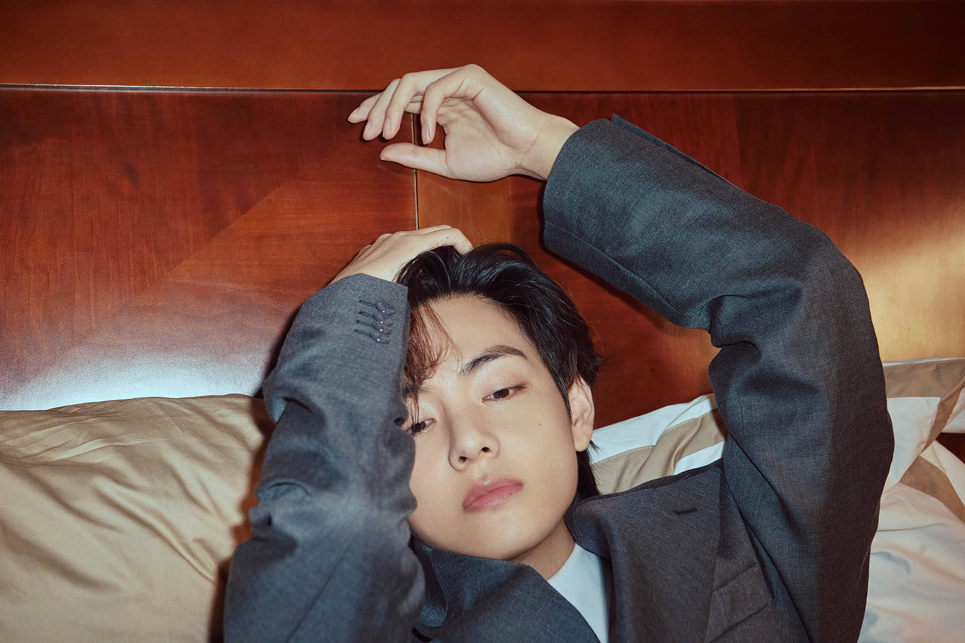 BTS V 床上休憩照 壁紙 — 一位身穿灰色西裝的年輕男子躺在木床頭板的床上，雙臂舉起，平靜地向上凝視。