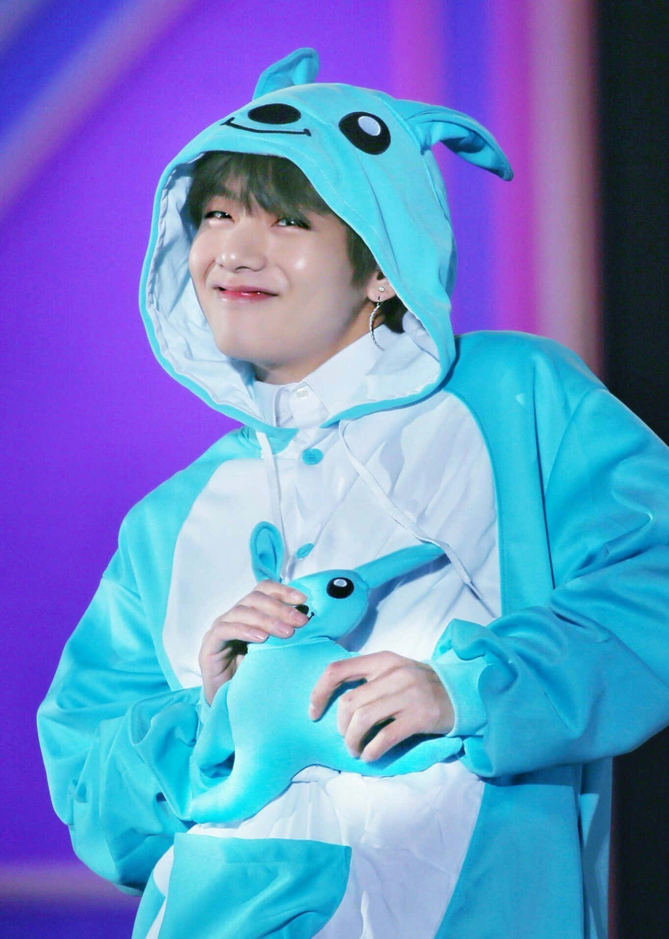 BTS V 展現迷人微笑 壁紙 — 一位穿著淺藍色兔子連體衣的表演者，在充滿活力的紫色舞台背景下，溫暖地微笑著，同時抱著一個相配的毛絨玩具。