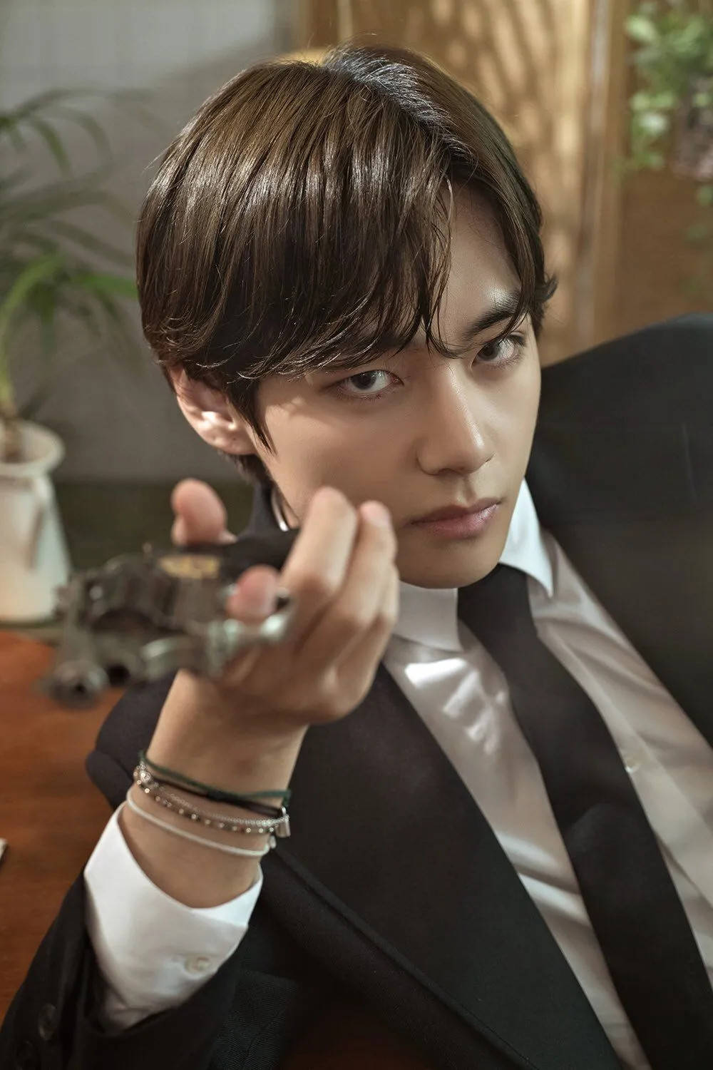 BTS V 持槍道具照 壁紙 — 一位身穿黑色西裝的年輕男子坐在一個光線溫暖、有植物和木質裝飾的房間裡，手持一部復古相機。