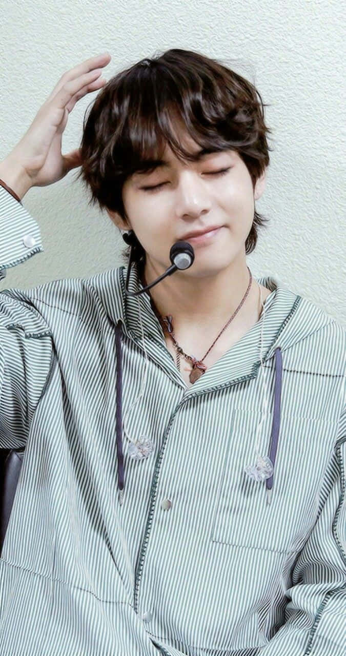 BTS V 散發真誠樂觀 壁紙 — 一位年輕男子，棕色亂髮，微笑著輕觸頭部，身穿條紋連帽衫和耳機麥克風。