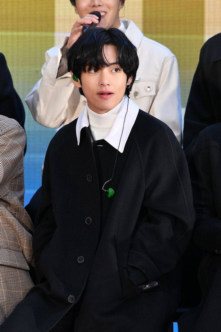 BTS V 時尚高領毛衣 壁紙 — 一位身穿黑色外套和白色高領毛衣的年輕男子，在明亮的黃色背景活動中專注地坐著。