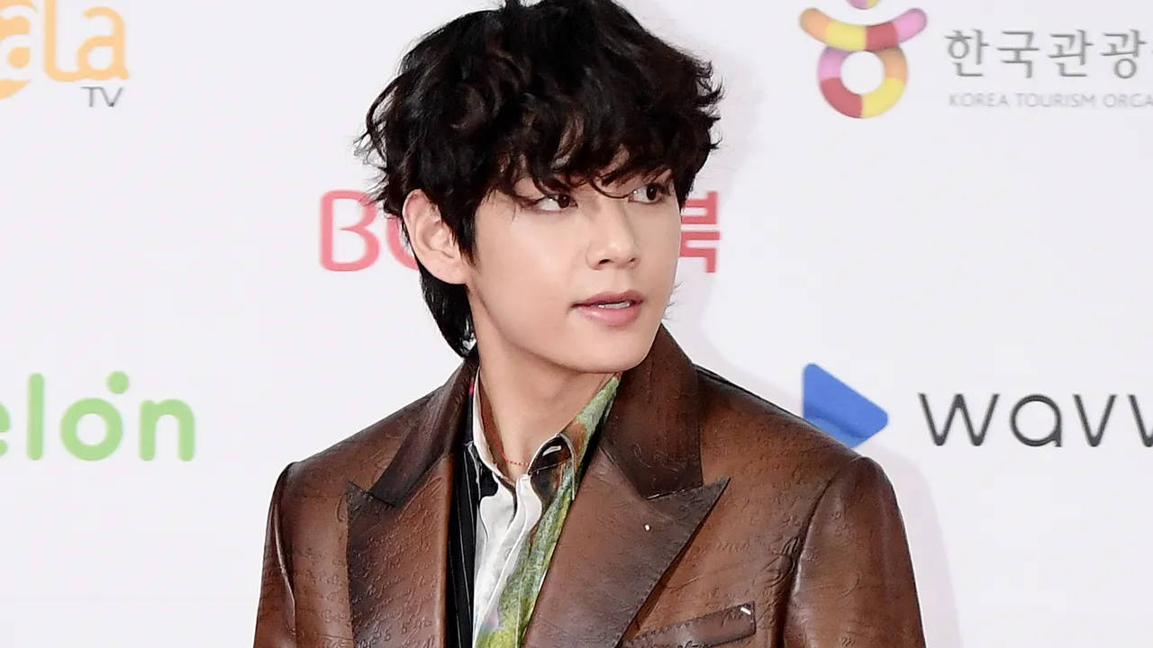 BTS V 棕色皮夾克 壁紙 — 一位深色捲髮的年輕男士在記者會上穿著一件質感豐富的棕色皮褸，內搭圖案襯衫。