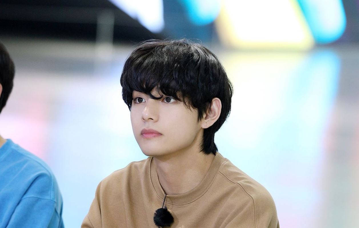 BTS V 棕色襯衫 壁紙 — 一位黑髮、身穿棕褐色襯衫的年輕男子，在模糊多彩的背景前若有所思地凝視。