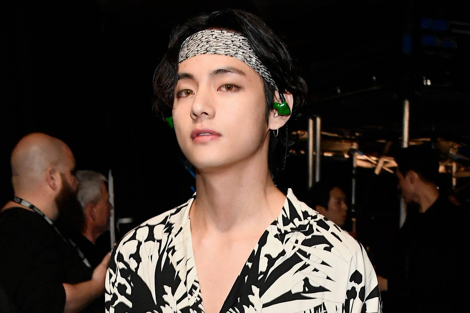 BTS V 極簡圖案服裝 壁紙 — 一位深色頭髮、戴著圖案頭帶的年輕男子，在舞台燈光下穿著大膽的黑白花卉襯衫。