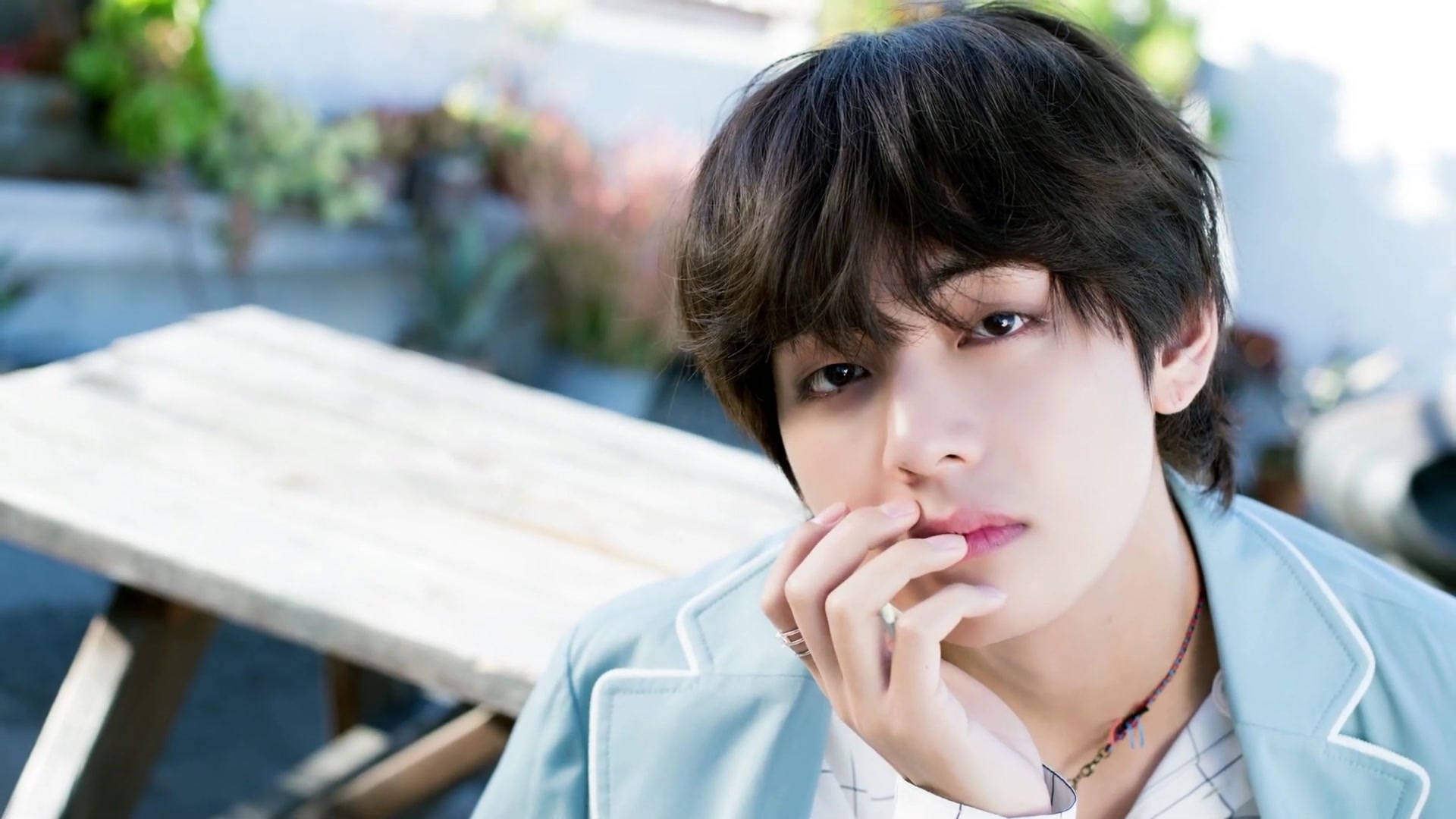 BTS V 浅藍色外套 壁紙 — 一位深色頭髮、身穿淺藍色外套的年輕男子，在木桌旁托腮沉思。