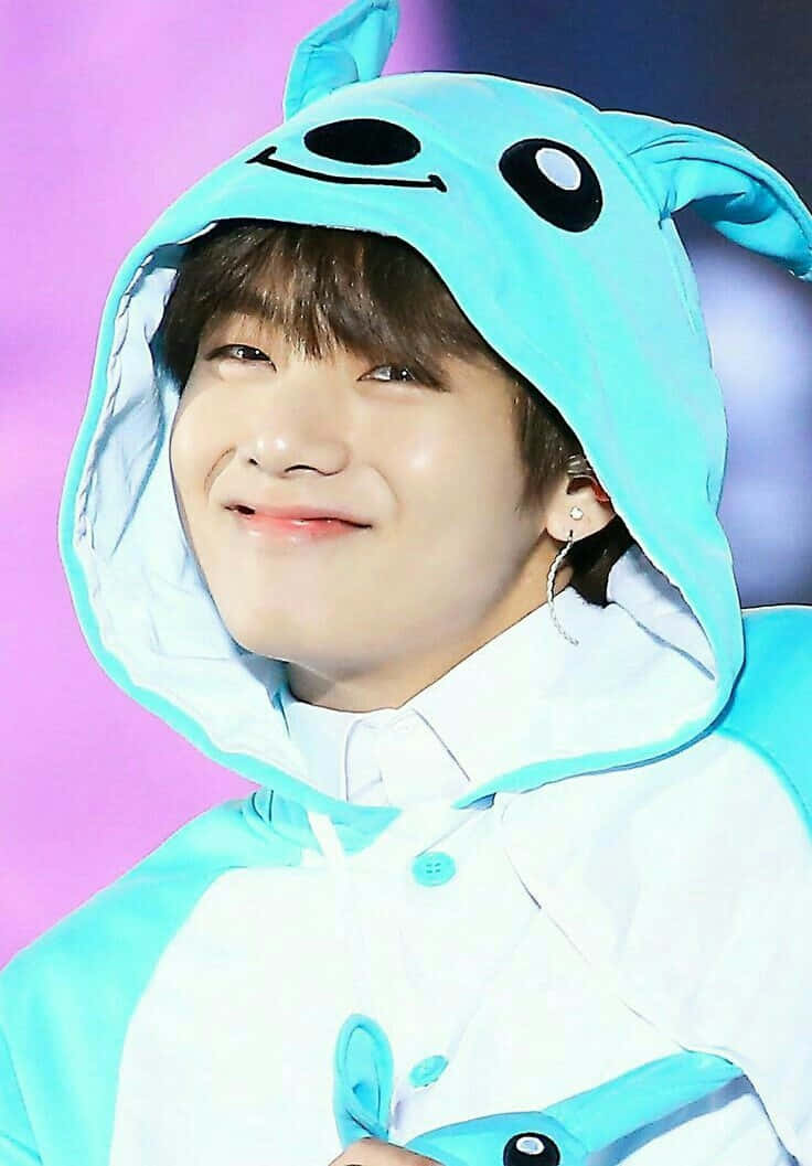 BTS V 為粉絲擺出俏皮姿勢 壁紙 — 一位年輕男子穿著可愛的淺藍色動物主題連帽服裝，在充滿活力的紫色背景下溫暖地微笑。