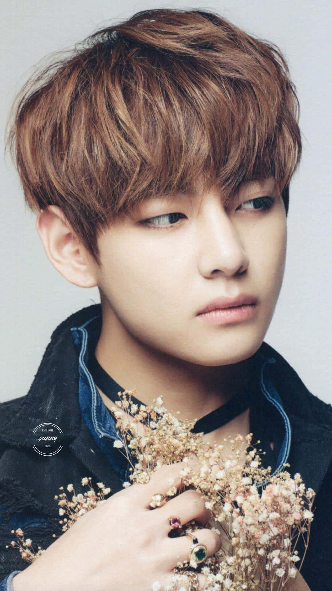 BTS V 無敵耀眼 壁紙 — 一位棕色亂髮的年輕男子若有所思地凝視著，手持乾花並戴著戒指。