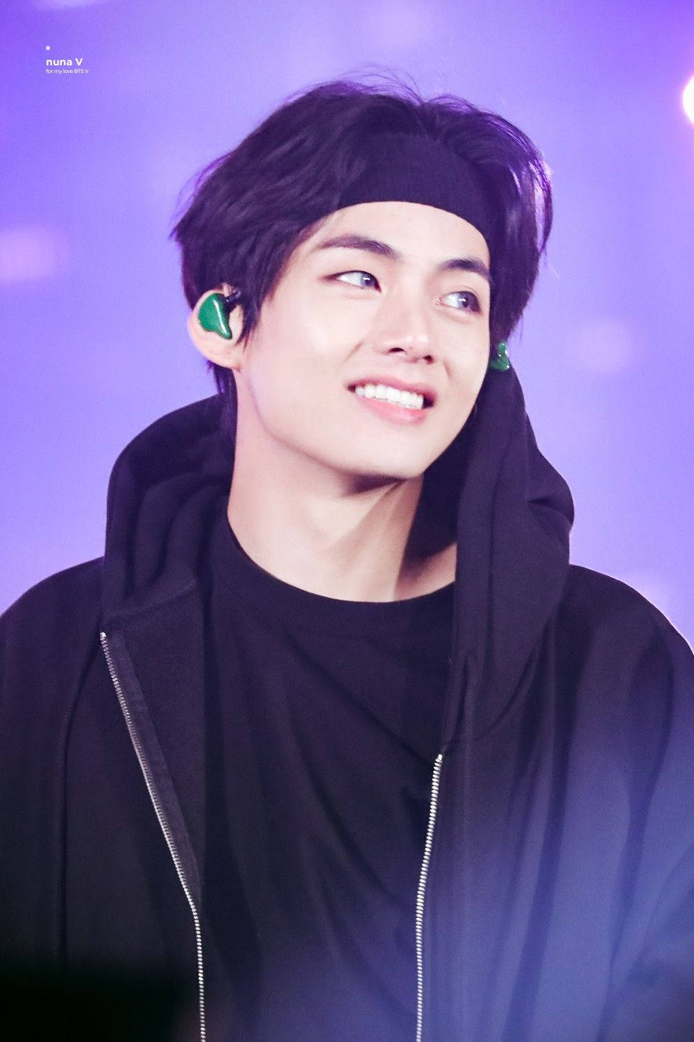 BTS V 時尚黑色造型 壁紙 — 一位黑髮、戴著綠色耳機的年輕男子，在發光的紫色舞台背景下溫暖地微笑。