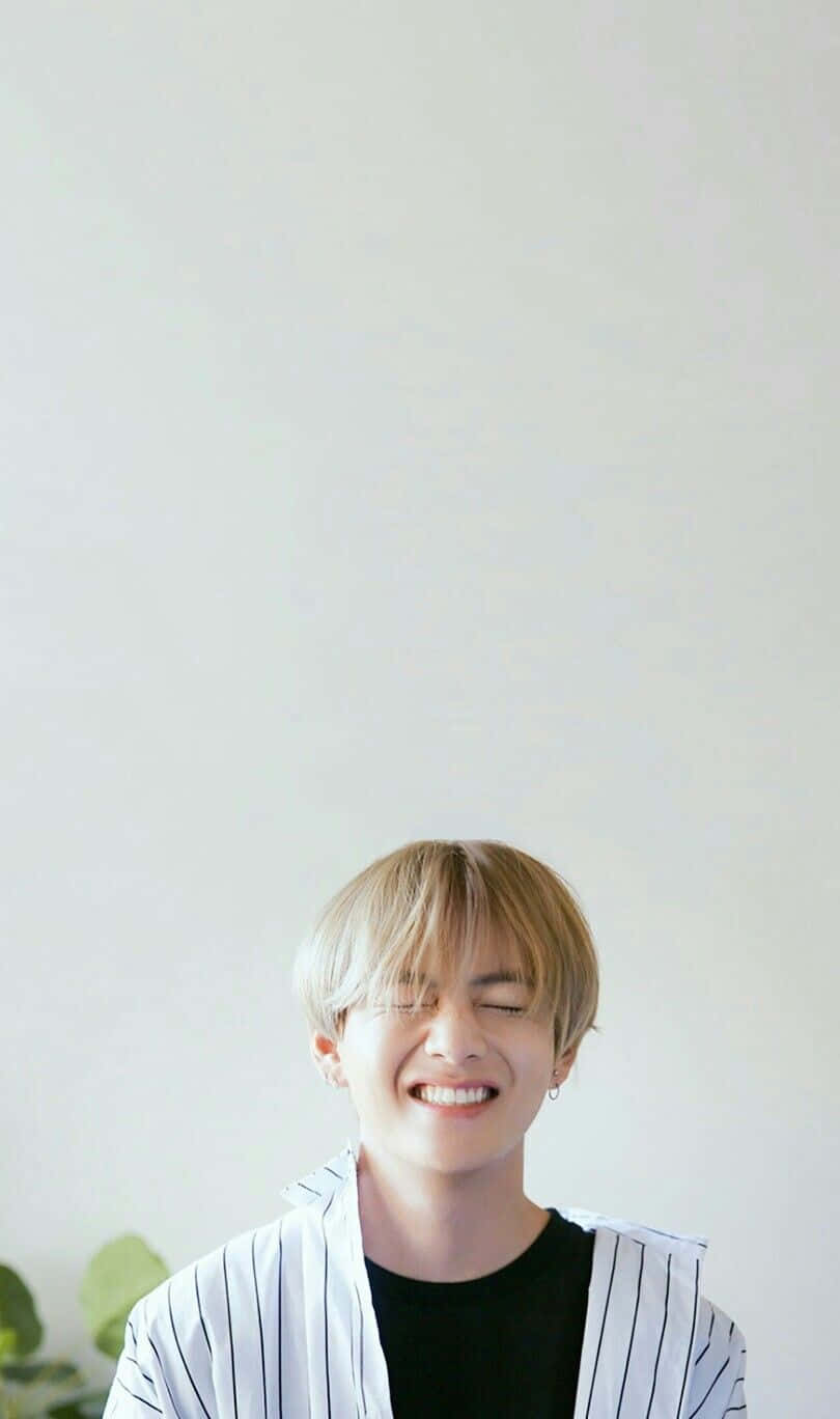 BTS V 站立於都市天際線前 壁紙 — 一位金髮年輕男子對著一面素白牆壁開懷大笑，牆腳有一株綠色植物探出頭來。