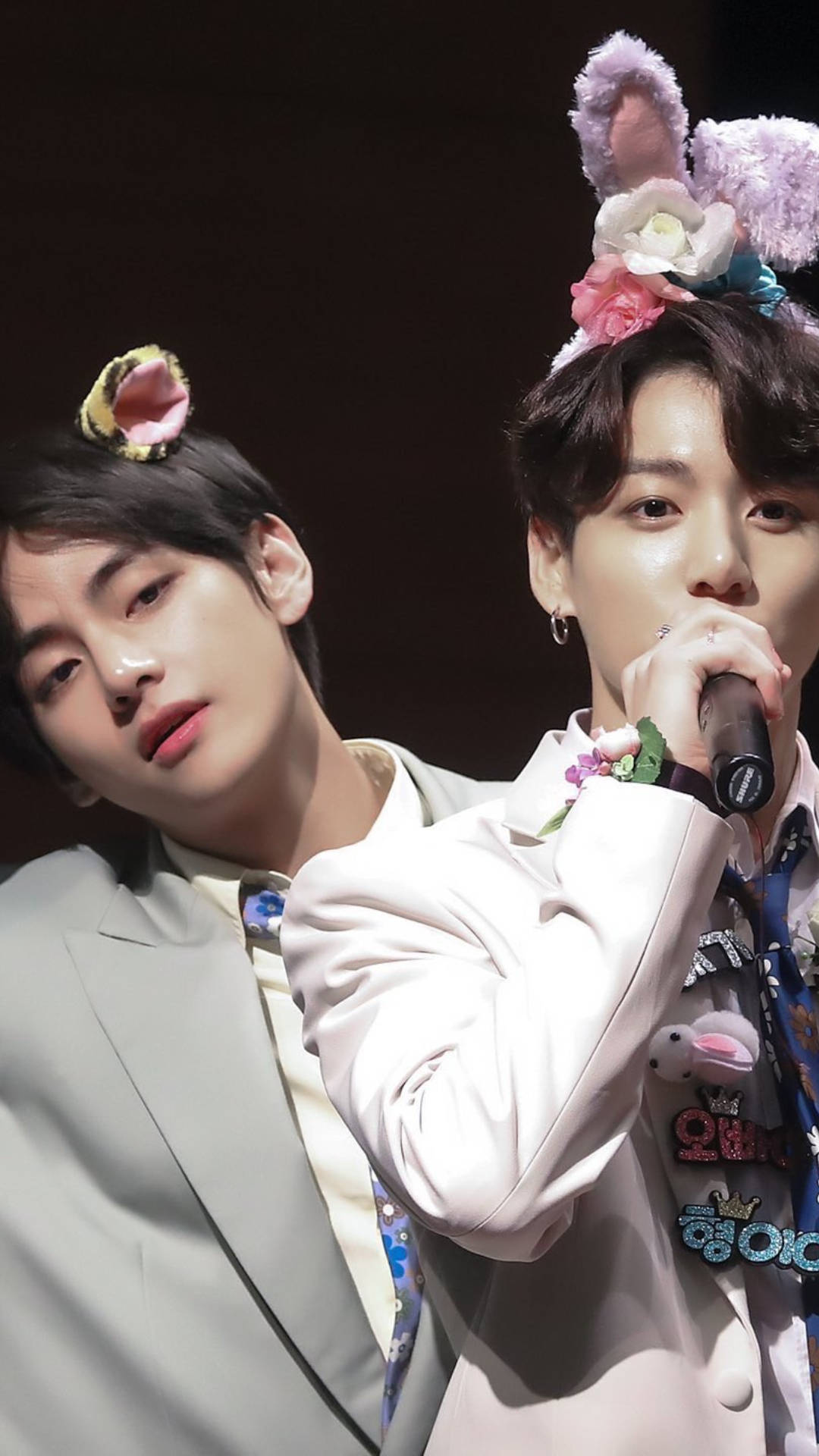 BTS V 與 Jungkook 戴可愛髮帶 壁紙 — 兩位身穿淺色西裝的年輕男子戴著花卉兔耳，其中一人在舞台燈光下對著咪高峰唱歌。