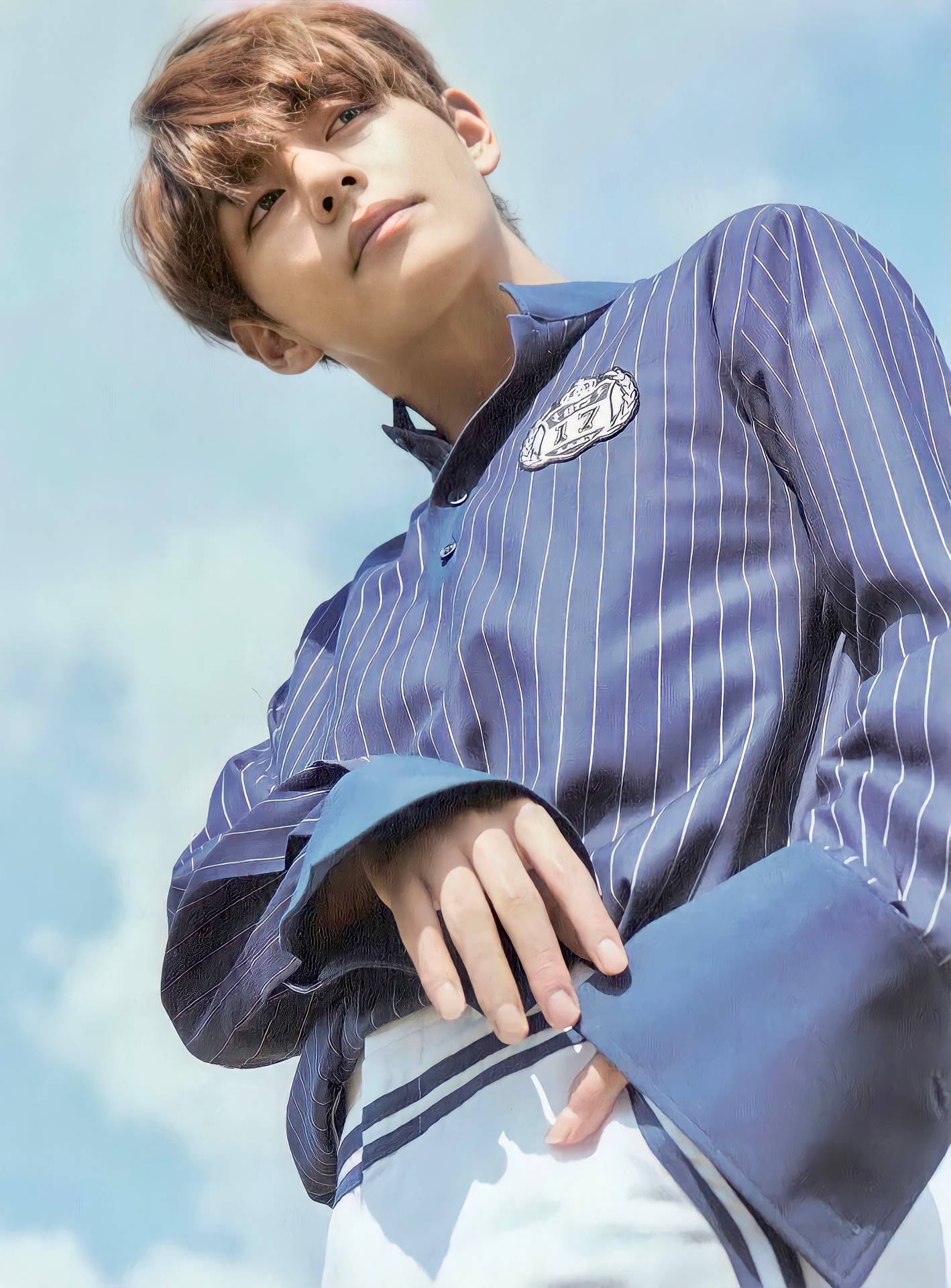 BTS V 藍色條紋襯衫 壁紙 — 一位身穿藍色條紋襯衫的年輕男子，在柔和雲朵的明亮天空下向上凝視。