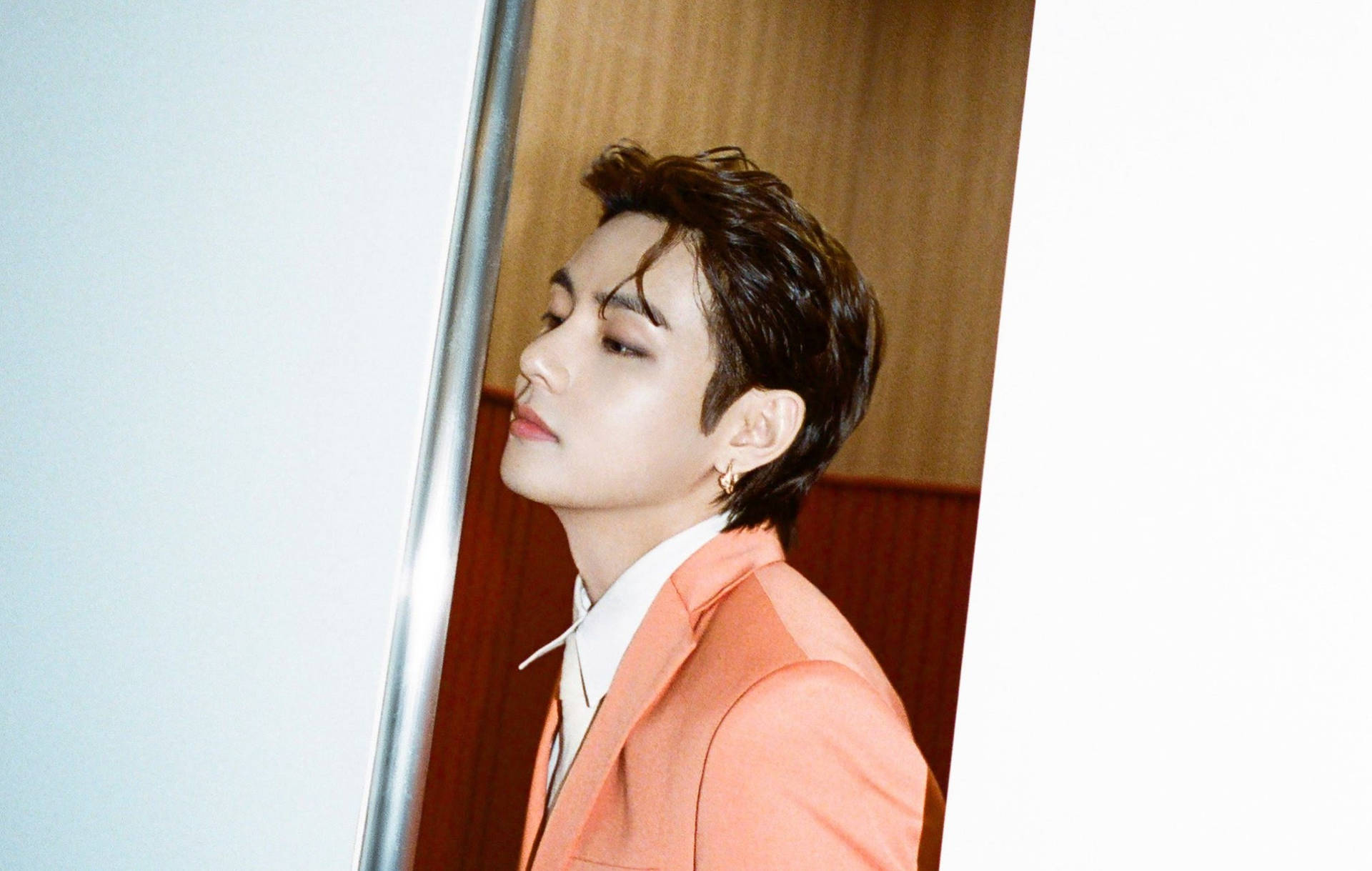 BTS V 頭髮向後梳 壁紙 — 一位身穿珊瑚色西裝的年輕男子在柔和光線的室內環境中閉著眼睛靠在牆上。