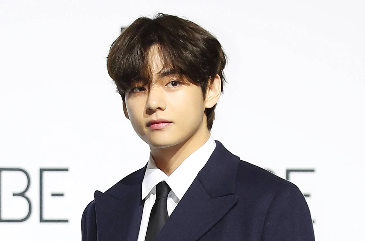 BTS V 黑色禮服造型 壁紙 — 一位深色頭髮的年輕男士穿著俐落的海軍藍西裝和黑色領帶，背景是純白色。