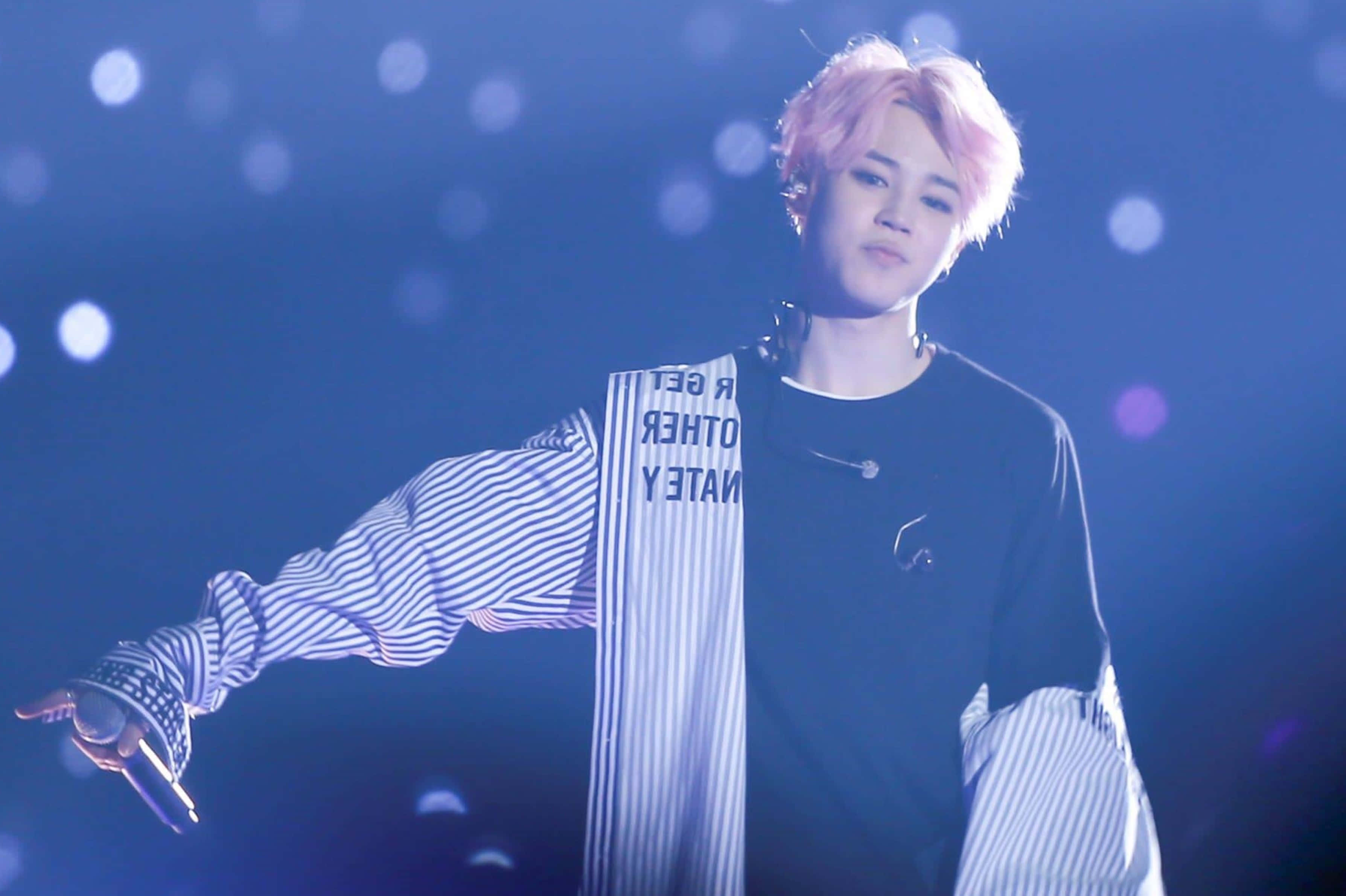 BTS Wings 巡迴演唱會 金珍洙 ARMY 拍攝高清快照 壁紙 — 一位粉紅色頭髮、條紋袖子的表演者在藍色燈光、散景燈光的舞台上做手勢。