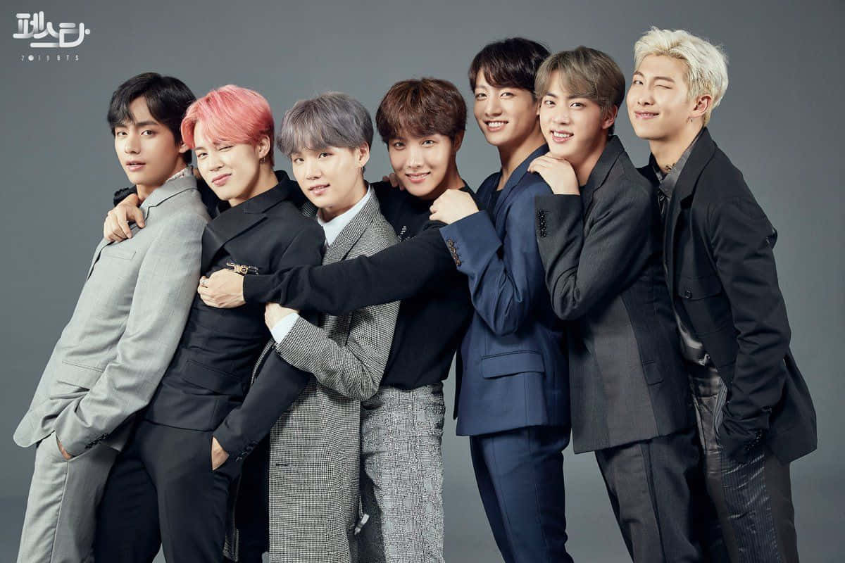 BTS x Mac 化妝大改造 壁紙 — 七位時尚的K-pop偶像穿著訂製西裝，在純灰色攝影棚背景前緊密擺姿勢，展現自信笑容和各種髮色。