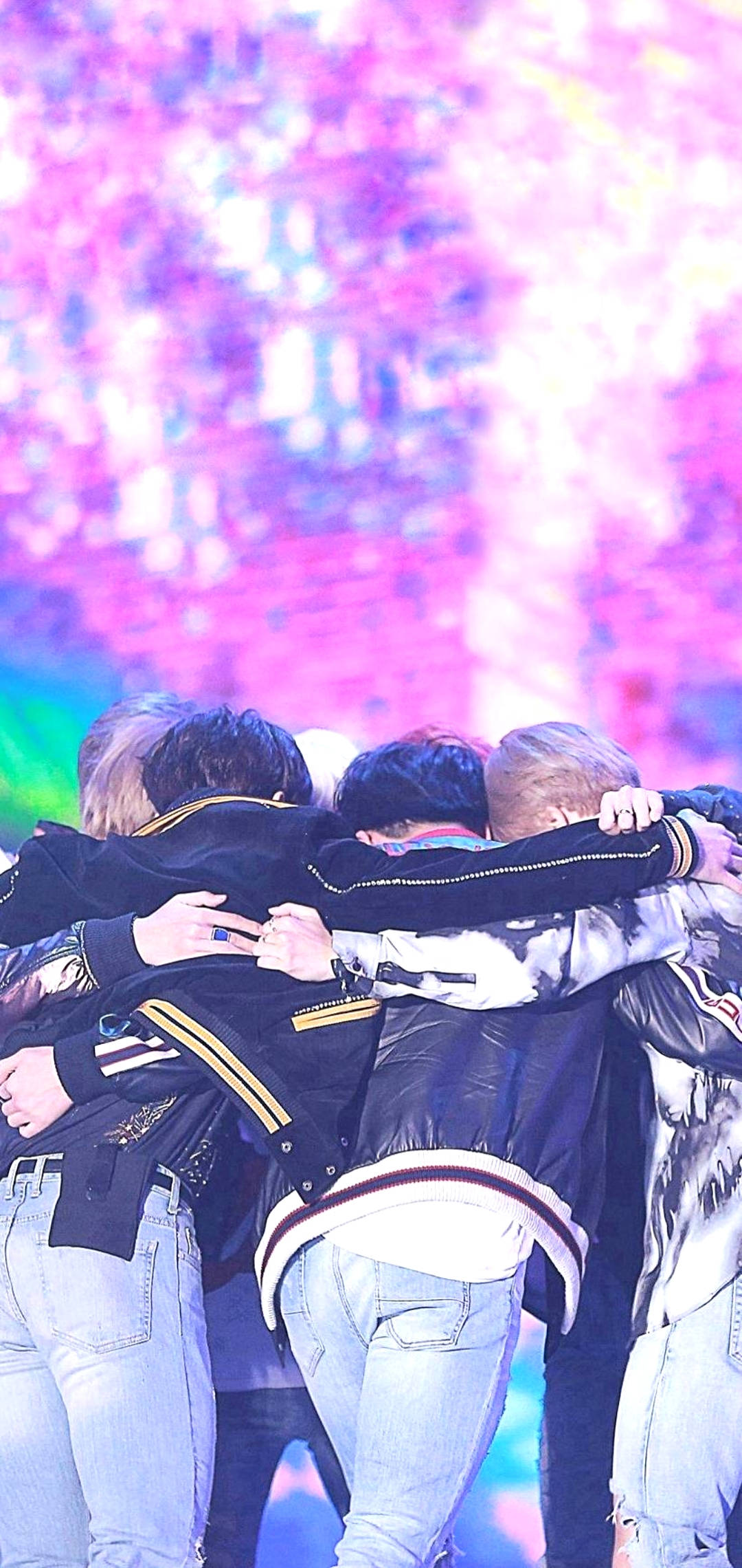 BTS 鎖定畫面 群體擁抱 壁紙 — 五個身穿外套的年輕男子擠在一個燈光璀璨的舞台上，身後是充滿活力的粉紅色和紫色燈光。