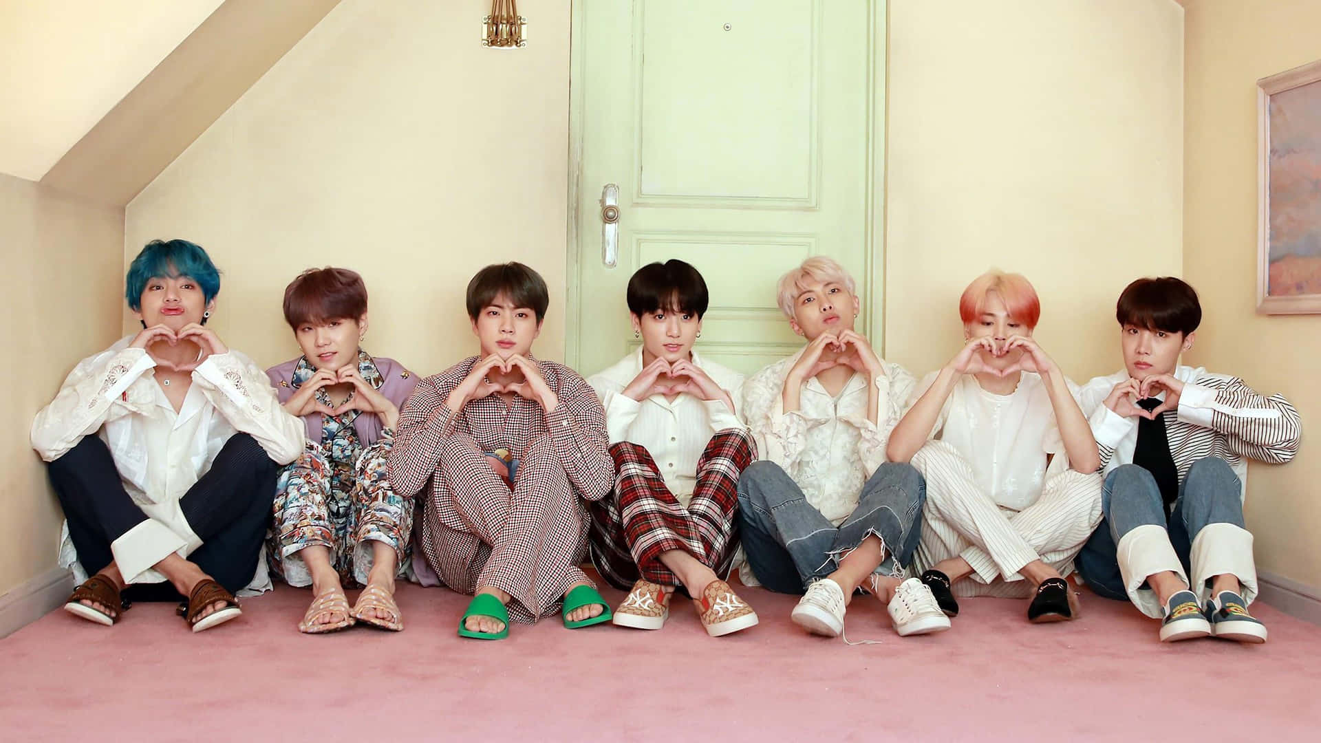BTS - 壁紙 — 七位擁有繽紛髮色的年輕男性在舒適的閣樓房間裡，排排坐在粉紅色地毯上，用手比出心形。