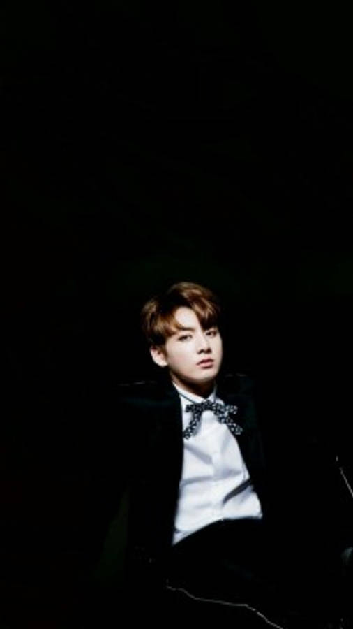 BTS 可愛風格：Jungkook 身穿黑色西裝 壁紙 — 一位身穿黑色西裝和領結的年輕男子在黑暗背景下抬頭仰望，帶有戲劇性的聚光燈效果。