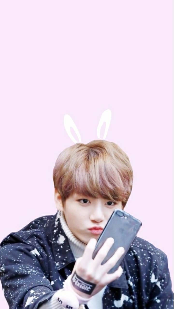 BTS 可愛風格：Jungkook 戴兔耳造型 壁紙 — 一位戴著兔耳朵的年輕男子在柔和的粉紅色背景下穿著星星夾克自拍。