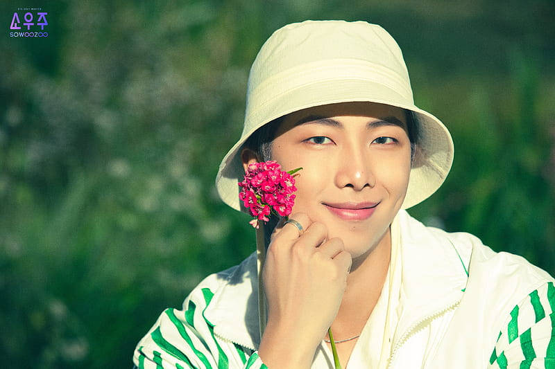 BTS 可愛風格：RM 手持粉紅花朵 壁紙 — 一位戴著白色漁夫帽的年輕男子，在茂密的綠色背景下，將一朵粉紅色的花靠近臉龐，溫柔地微笑。