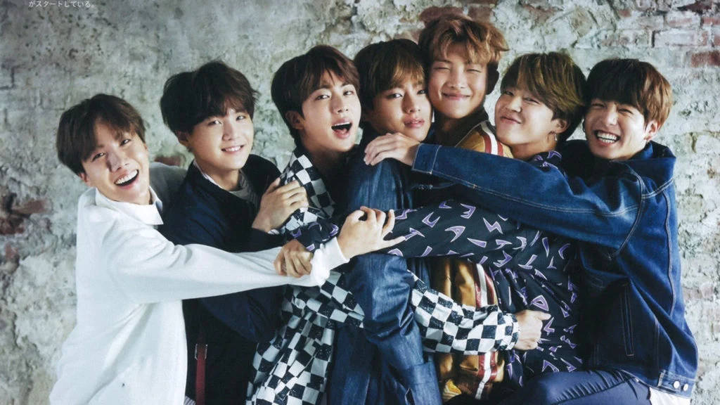 BTS 可愛風格：擁抱群像 壁紙 — 七個微笑的年輕男子穿著休閒服裝，擠在一起靠著一面風化的磚牆，表情歡樂。