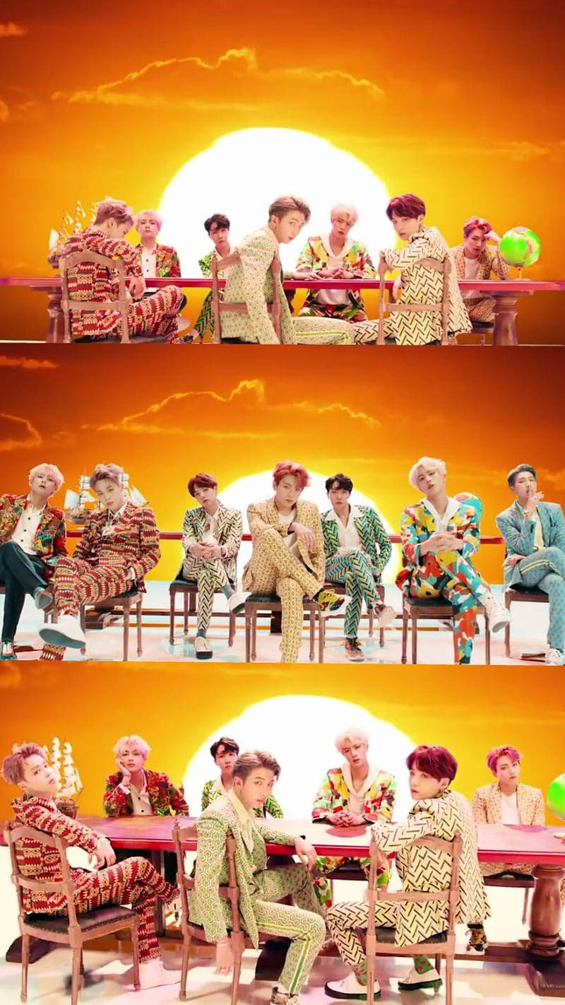 BTS 可愛風格：日落時分休憩 壁紙 — 七個穿著色彩繽紛圖案西裝的男人圍坐在一張紅色桌子旁，背景是充滿活力的橙色日落和一個巨大的太陽。