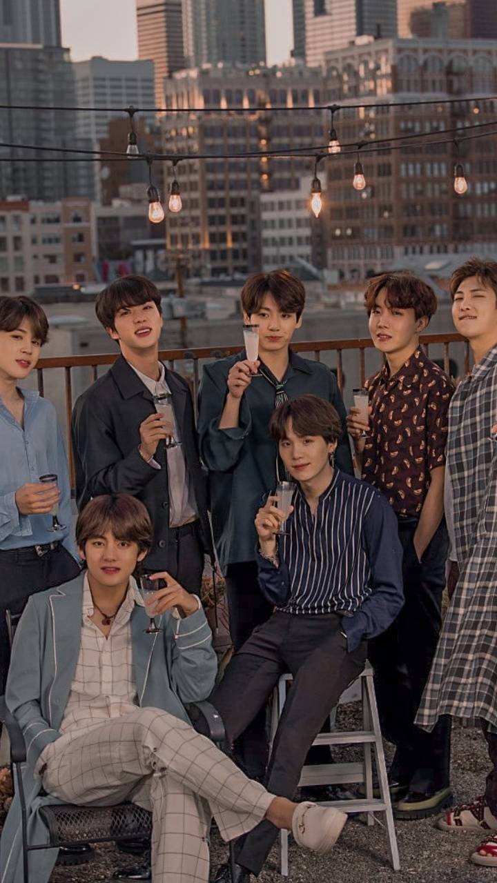 BTS 喝酒美學壁紙 壁紙 — 七位穿著時尚的年輕男子在屋頂上放鬆，背景是城市天際線，在黃金時刻燈串閃爍。