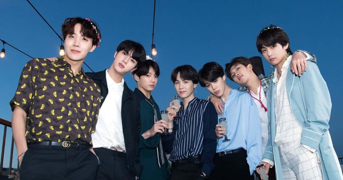 BTS 夜空藍色主題照 壁紙 — 七位時尚年輕男子在黃昏時分於天台一起擺姿勢，在串燈下舉杯，背景是深藍色的天空。