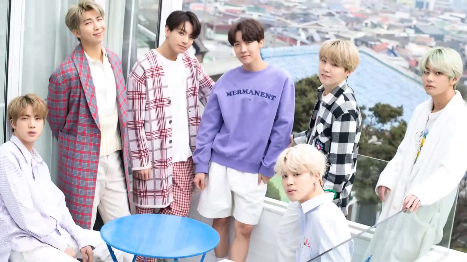 BTS 夜間陽台休閒照 壁紙 — 七位時尚的年輕男士穿著休閒格仔和粉彩服裝，在俯瞰廣闊城市景觀的玻璃陽台上自信地擺姿勢。
