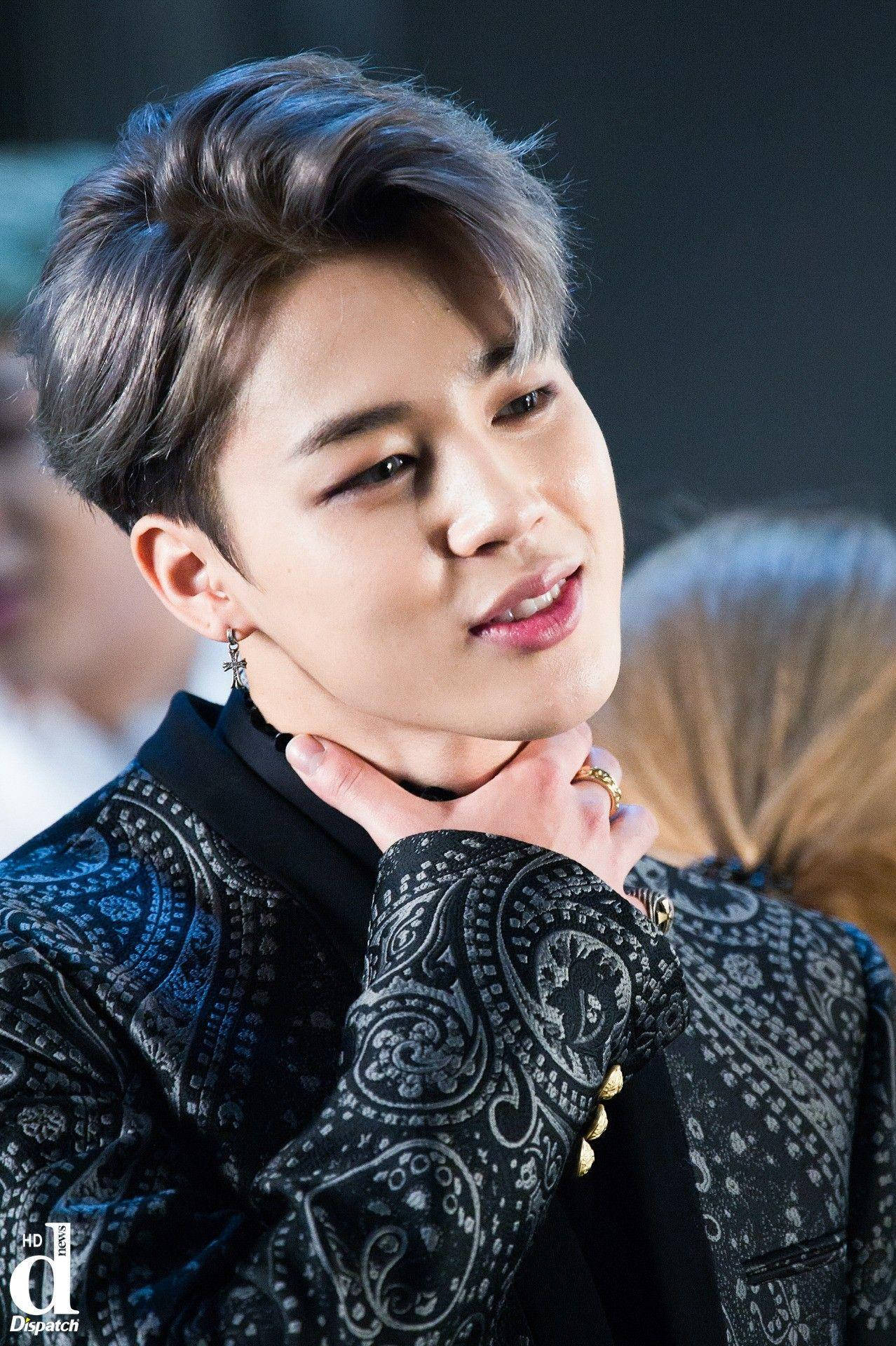 BTS 崔勝哲 可愛正式風 壁紙 — 一位年輕男子，留著時尚髮型並戴著耳環，穿著帶有圖案的黑色外套，輕輕觸摸自己的頸部。