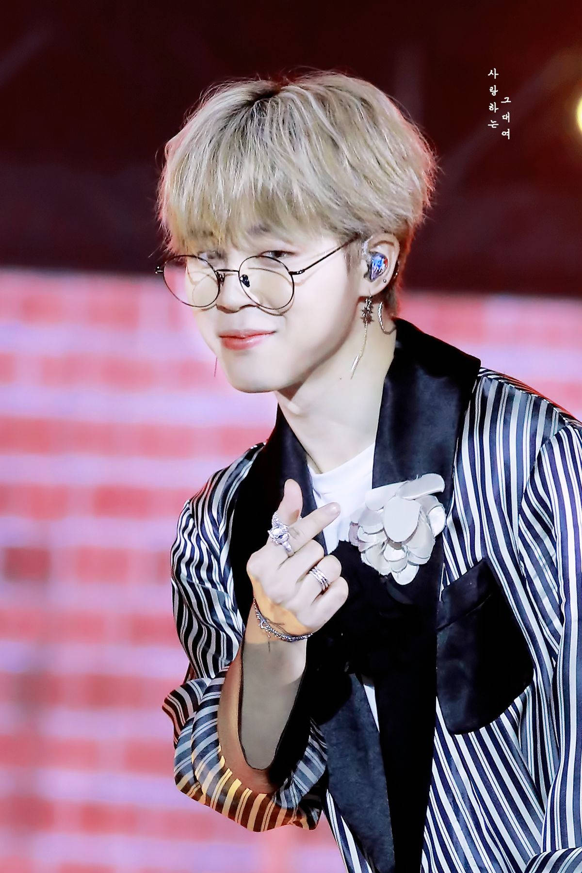 BTS 崔勝哲可愛手指心形 壁紙 — 一位金髮戴眼鏡的年輕男子在明亮燈光下的紅磚舞台上微笑比出和平手勢。