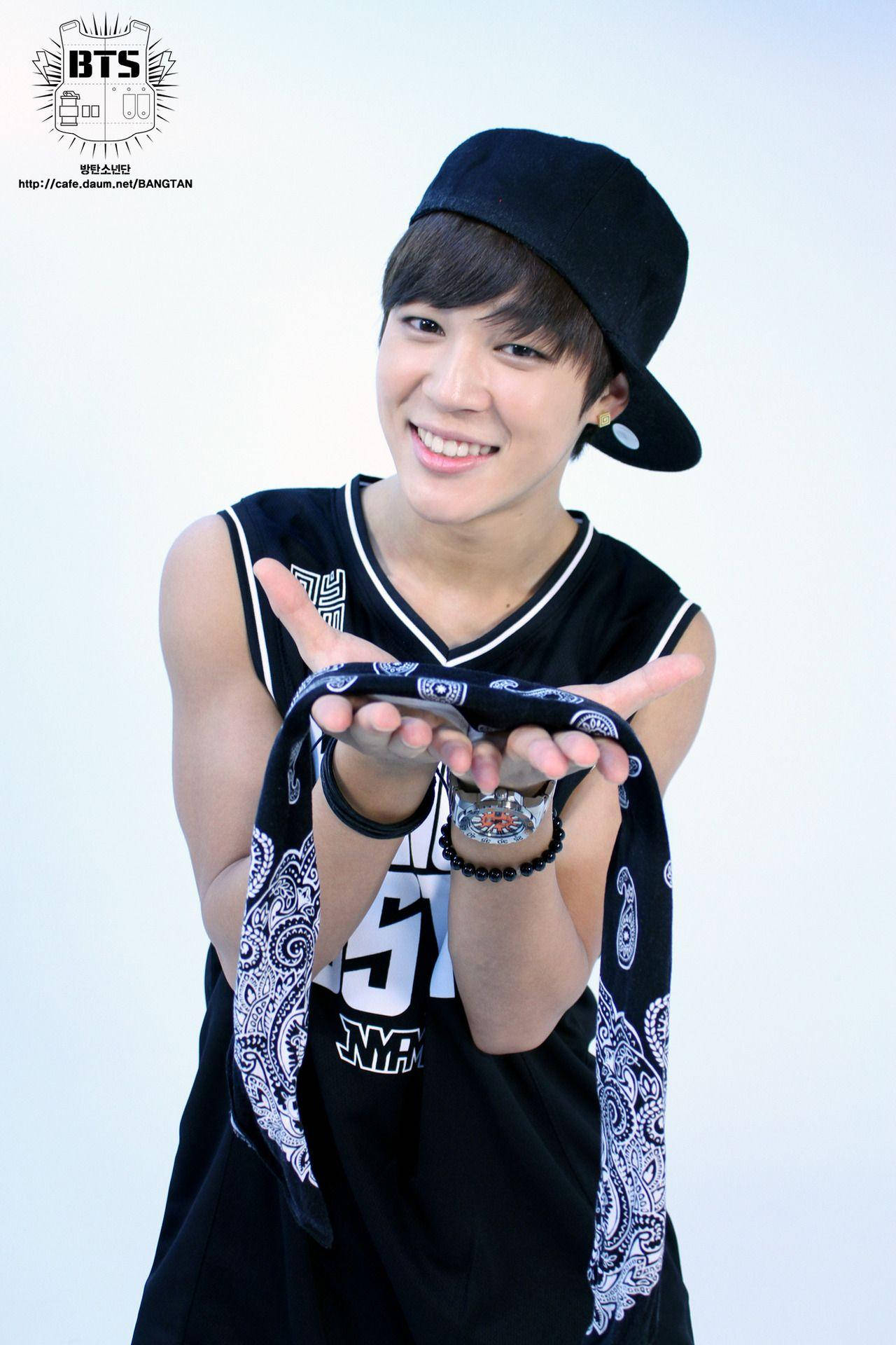 BTS 崔勝哲可愛頭巾 壁紙 — 一位微笑的年輕男子，戴著黑色鴨舌帽和無袖運動衫，在純白色背景前，張開手掌，拿著一條圖案頭巾。