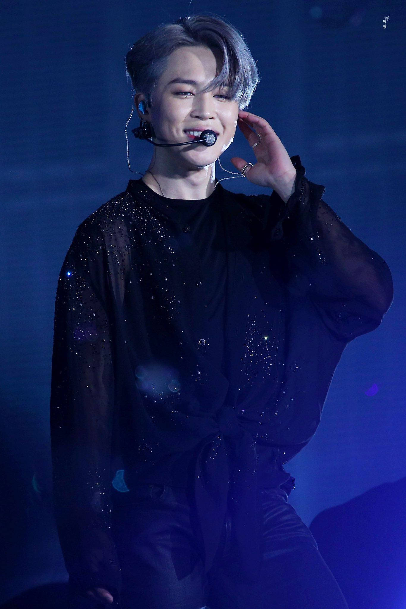 BTS 崔勝哲黑色時尚可愛 壁紙 — 一位面帶微笑、銀色頭髮的表演者，穿著閃亮的黑色服裝，在藍色舞台燈光下對著麥克風唱歌。