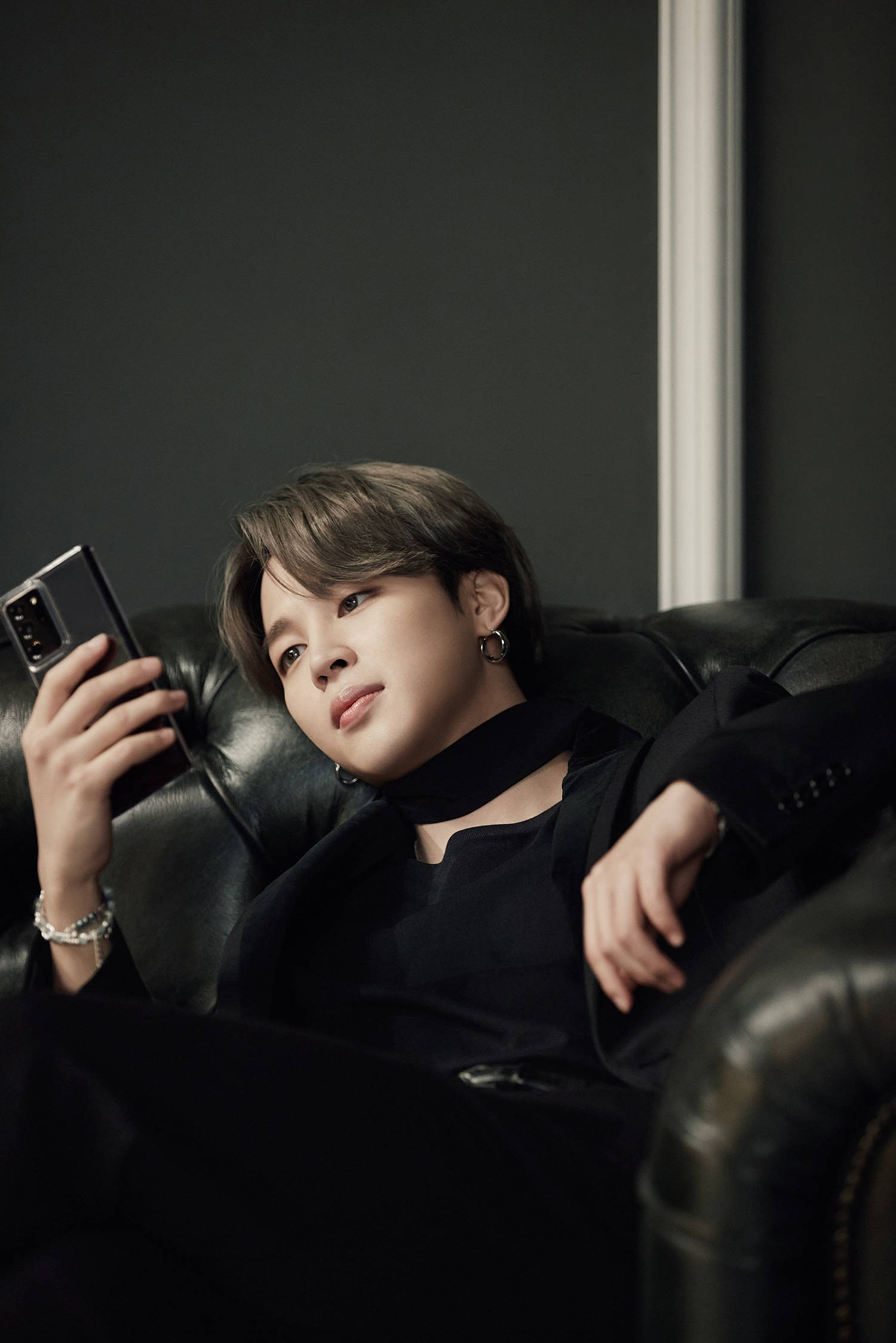 BTS 崔然竣 Samsung 品牌代言 壁紙 — 一名全身黑衣的年輕男子懶洋洋地躺在皮梳化上，手持電話，背景柔和模糊。