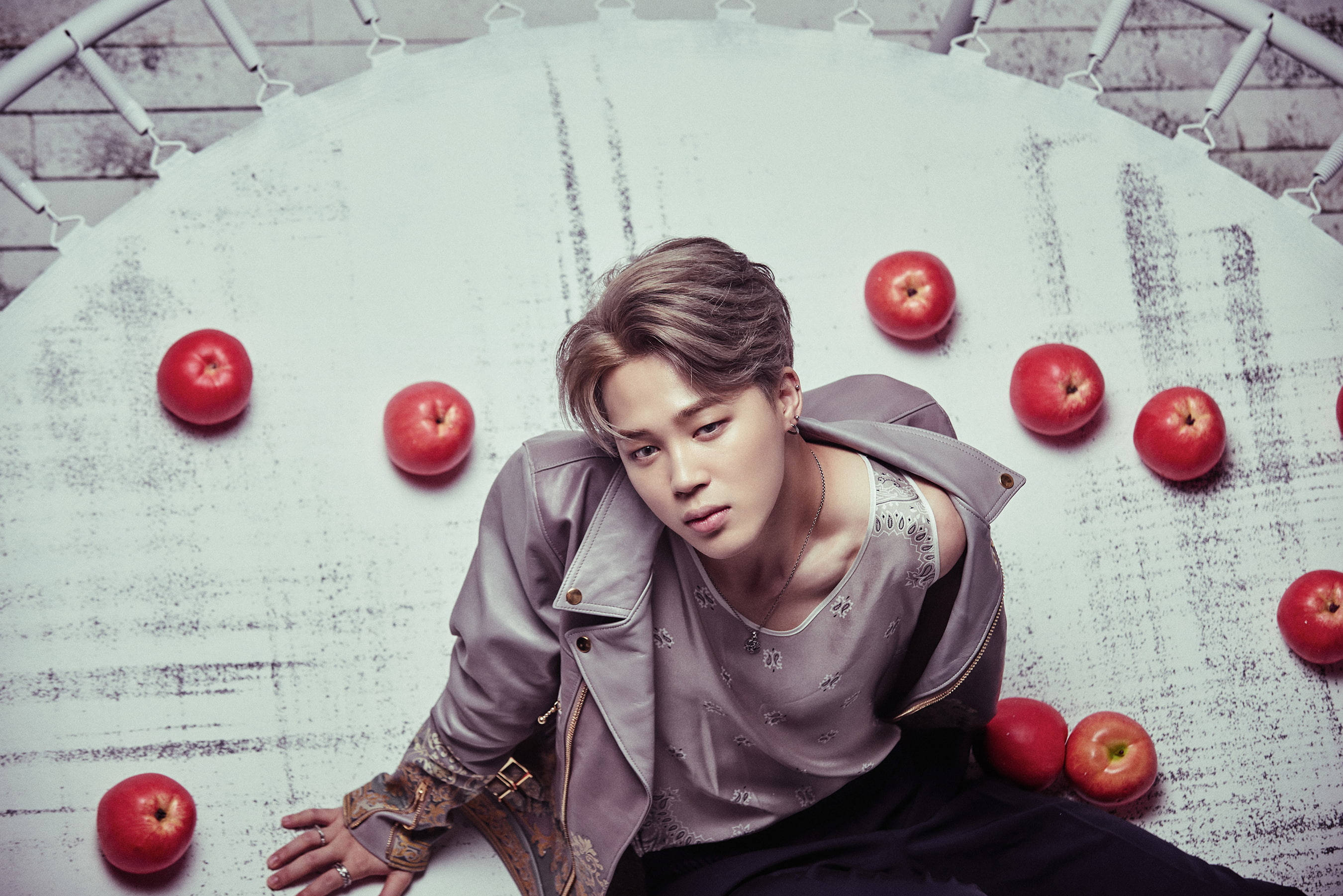 BTS 崔然竣 Wings 壁紙 — 一位身穿灰色外套的年輕男子坐在一個磨損的白色表面上，周圍是紅蘋果，頭頂有一個金屬框架。